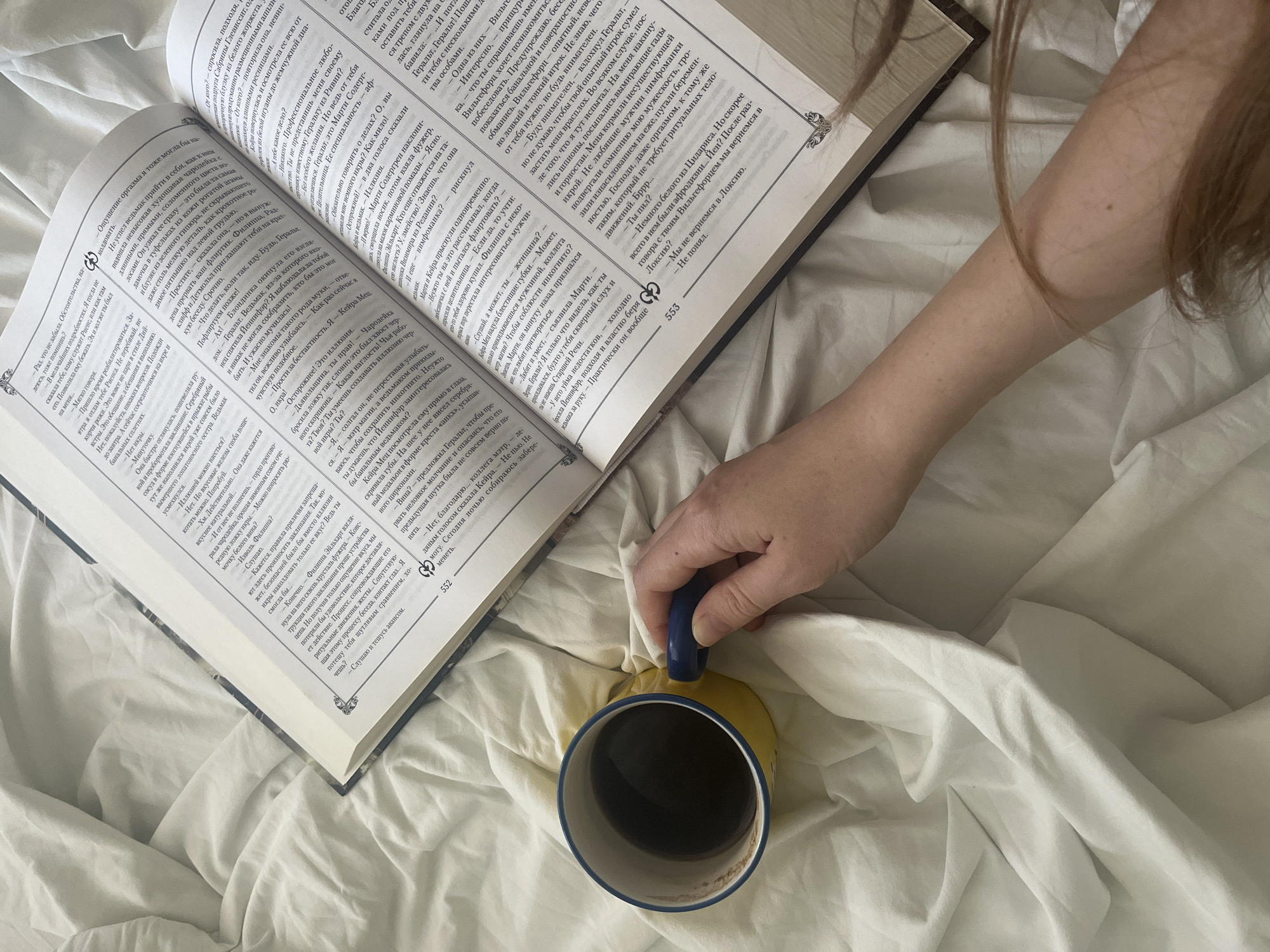 The image depicts a still life scene, likely indoors on a bed covered with white sheets. The primary subjects are an open book and a yellow mug filled with a dark liquid, presumably coffee or tea. A person's hand, with a blue ring on the index finger, is visible reaching towards the mug. The book is open to pages with text, and the person's arm and hair are also partially visible, suggesting they are either reading or resting. The overall impression is one of quiet relaxation or leisure. There is no identifiable information to place this scene in Komsomolskoye, Ukraine.