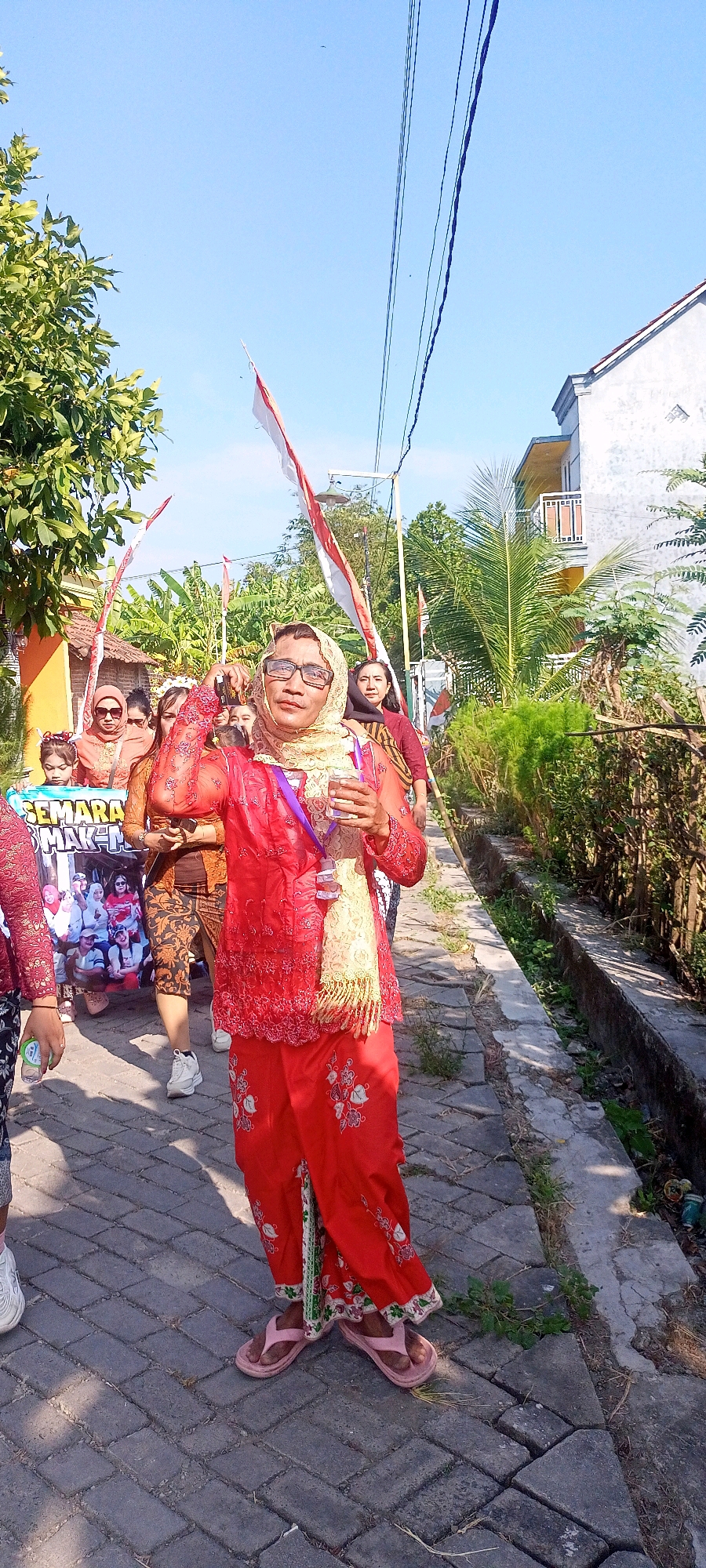 In the city of Gempolklutuk, Indonesia, a group of people are participating in a parade. The foreground is dominated by a person wearing a bright red outfit, a headscarf, and glasses, holding a glass in their right hand. They are walking on a brick-paved path. Behind them, other participants are visible, including some holding Indonesian flags. A banner with an image of people is also being carried. The environment features trees, houses, and a drainage ditch along the side of the path. The sky is clear and blue, indicating it is a sunny day.