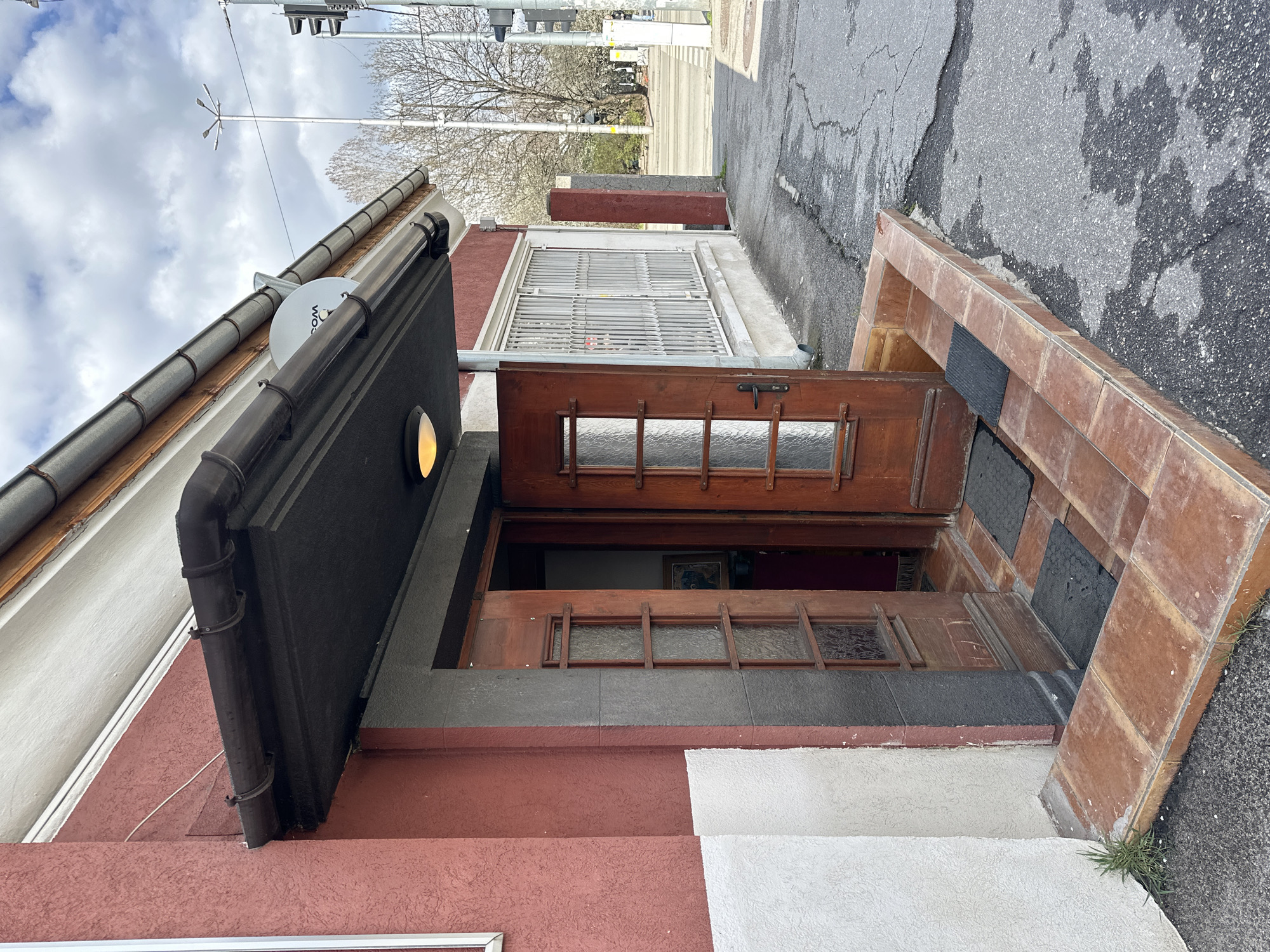 Here is a detailed description of the image:
The image presents an unusual perspective of a building facade in Sofia, Bulgaria, likely taken from a low angle and then rotated. The primary focus is the entrance of a structure, with an open wooden door featuring glass panes. The door is set within a frame, with a similar frame above it, partially obscured by a window with metal bars.
The building’s facade is painted in sections with different colors: red, white, and dark gray. Gutters and drainpipes are attached to the building. A satellite dish is visible on the roof. Above the entrance, a lamp is mounted, emitting a yellow glow.
The entryway consists of brickwork with inlaid dark tiles. The road nearby shows signs of wear and tear, with cracks and patches.
In the background, a street scene is visible, complete with traffic lights and trees. The sky is overcast with some cloud cover. There are no people visible in the frame.