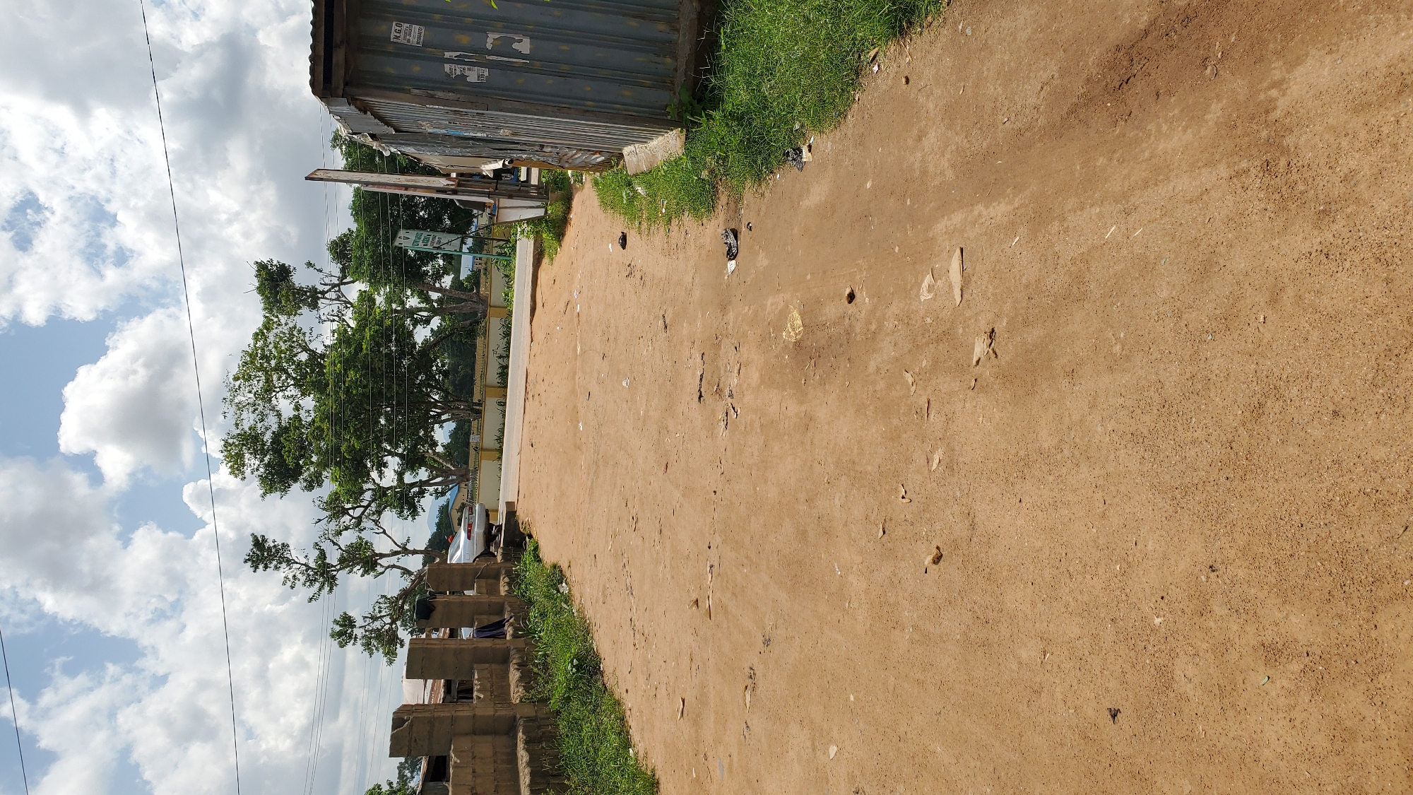 The image shows a dirt road in Jalingo, Nigeria. The road is surrounded by vegetation and some buildings. There is a car parked on the side of the road, and a few people can be seen in the distance. The sky is cloudy, and the sun is shining. The scene appears to be a typical street in Jalingo, a city located in northeastern Nigeria, known for its dusty roads and green vegetation. The image captures a moment of everyday life in this Nigerian city.