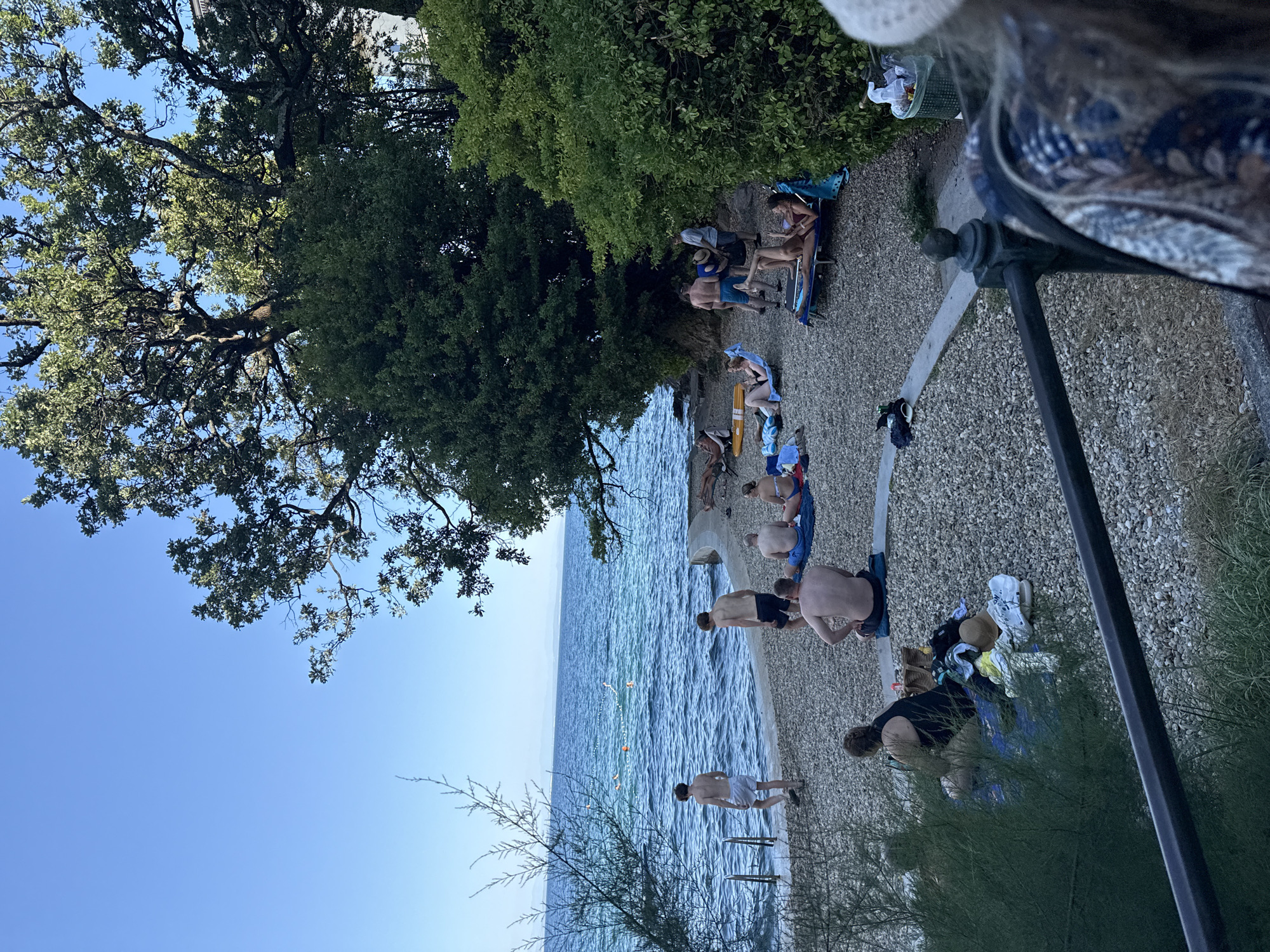 This is a photograph of a rocky beach with several people enjoying the water and sun. The scene appears to be located in Grad Opatija, Croatia, given the distinctive trees and coastal landscape. In the foreground, a person wearing a white hat and patterned clothing is partially visible, suggesting they are on a balcony or elevated viewing point. The beach is populated by several individuals in swimwear, some sitting on towels and beach chairs, while others are near the water's edge. Two young men are standing by the water, with one walking towards the sea. In the water, a buoys create a marked swimming area. The background features lush green trees, a clear blue sky, and distant hills. The overall ambiance is one of a relaxed summer day at the seaside.