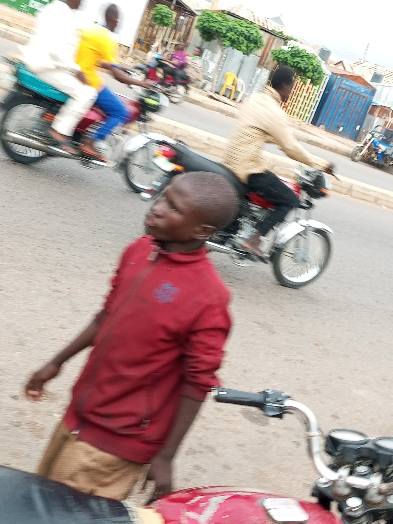 A young boy, wearing a red zipper jacket and tan pants, is in the foreground, looking to his left. He is partially obscured by what appears to be the handlebar and front of a motorcycle, also red. In the background, two individuals are riding a motorcycle. The person in front is wearing a yellow shirt and blue pants, while the person behind them is wearing a white robe. The motorcycle is dark red and silver. Further in the background, another person on a motorcycle is visible, wearing a striped long-sleeved shirt and black pants. This scene is taking place on a paved road in Tempure, Nigeria. The environment suggests an urban setting with some buildings and greenery visible.