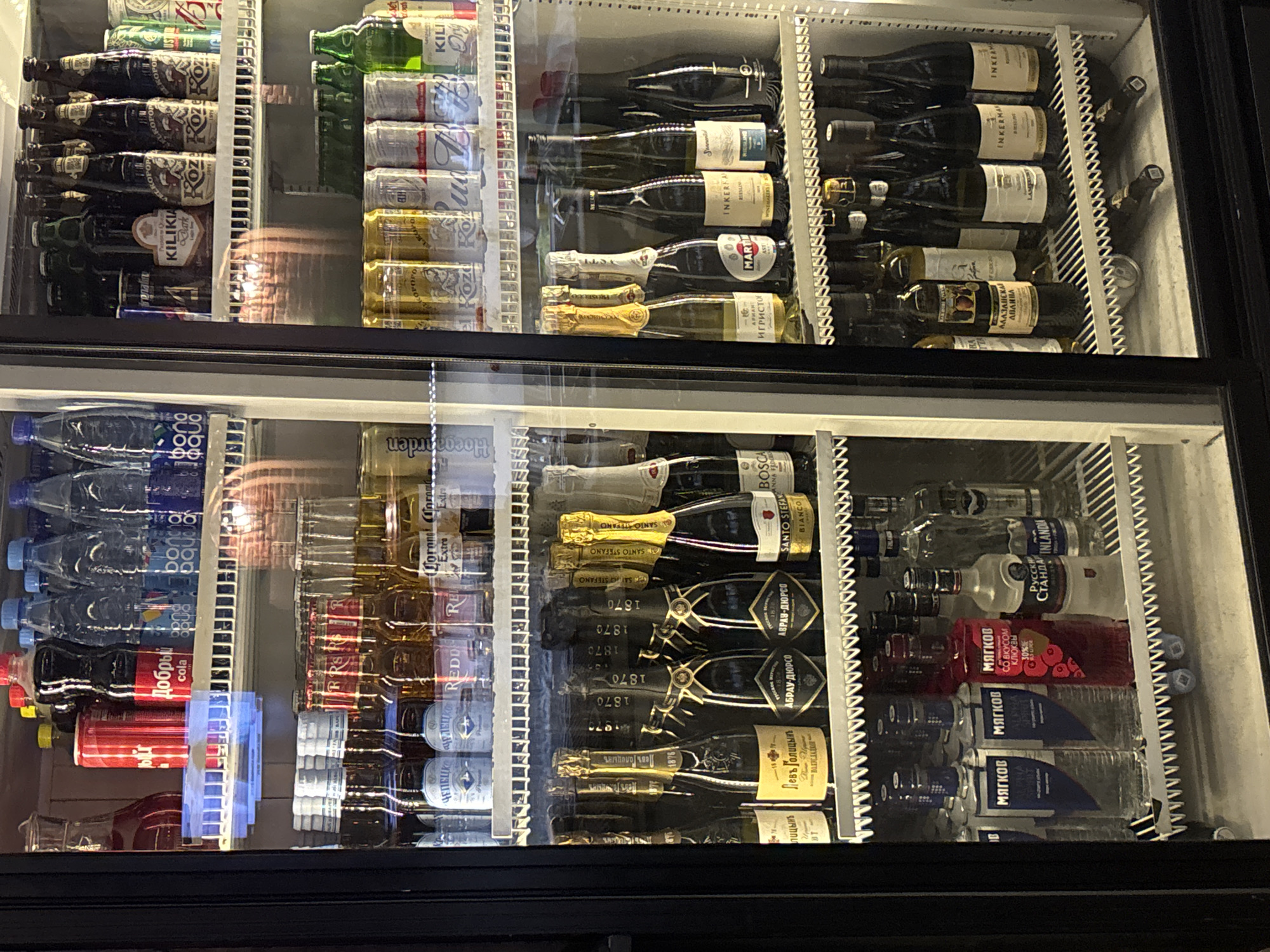 The image shows the inside of a commercial beverage refrigerator, likely a display cooler in a store or bar. The refrigerator is divided into two vertical sections by a central divider. The left section contains rows of glass bottles filled with various beverages, including what appear to be sodas, water, and possibly alcoholic drinks. Some bottles have prominent labels in Cyrillic script. There are also red cans visible on the top shelf. The right section contains a similar assortment of glass bottles and cans. Some bottles on the right are dark-colored, potentially wine or spirits, with labels also in Cyrillic.
The refrigerator shelves are made of white wire racks. The interior of the refrigerator appears well-lit, with reflections visible on the glass doors, suggesting an ambient indoor environment. The specific location in Chaikovsky, Russia, cannot be determined from the image alone. No people are present in the image, and there are no actions or interactions depicted. The scene is a static display of commercial beverages.