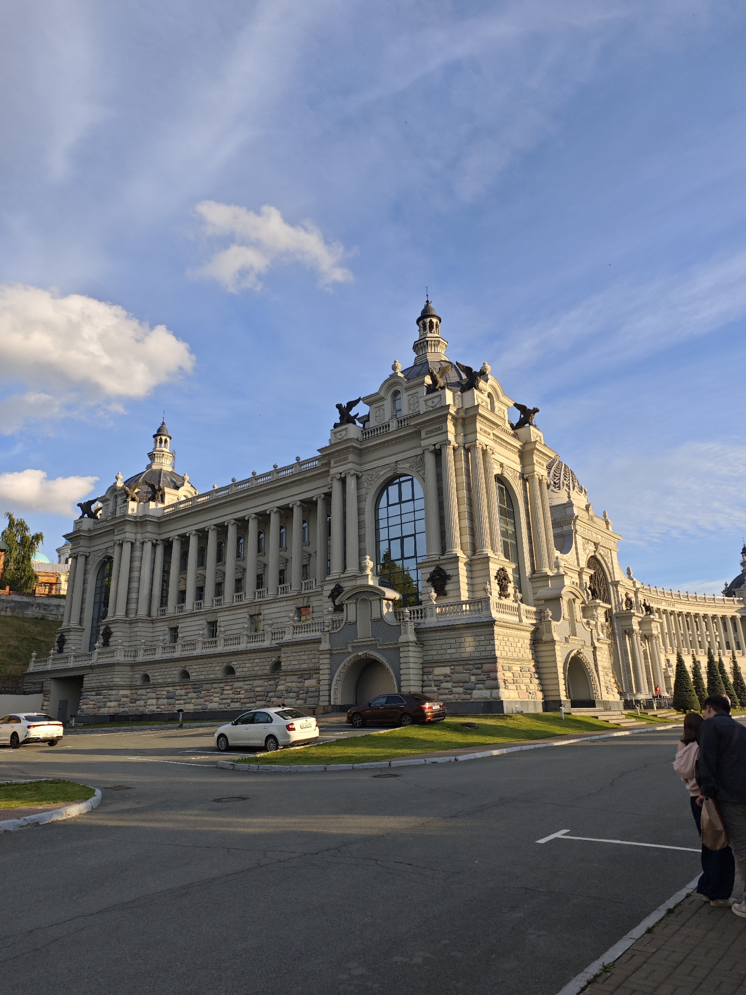 The image shows a grand, ornate building with neoclassical architecture under a bright blue sky with scattered clouds. The building features a prominent dome, numerous columns, arched entrances, and detailed sculptures. The sun is casting long shadows, suggesting it is late afternoon or early evening.
In the foreground, there is a paved area with a few cars parked. One white car is on the left, and another white car is parked on the grass to the right. A dark-colored car is partially visible near an arched entrance of the building. Two people, a man and a woman, are standing on the sidewalk in the lower right corner of the image, facing away from the camera, appearing to be observing the building or the surroundings. They are casually dressed.
The scene appears to be a public space, possibly a park or a plaza, with neatly manicured grass areas around the building. The sky is a vibrant blue with some wispy cirrus clouds and larger, fluffy cumulus clouds, indicating fair weather. The architectural style of the building, with its symmetry, classical elements, and imposing scale, suggests a historical or governmental significance. The light and shadows create a sense of depth and highlight the building's intricate facade. No visible text is present in the image. The location context is Kazan, Russia, and the building is likely a significant landmark.