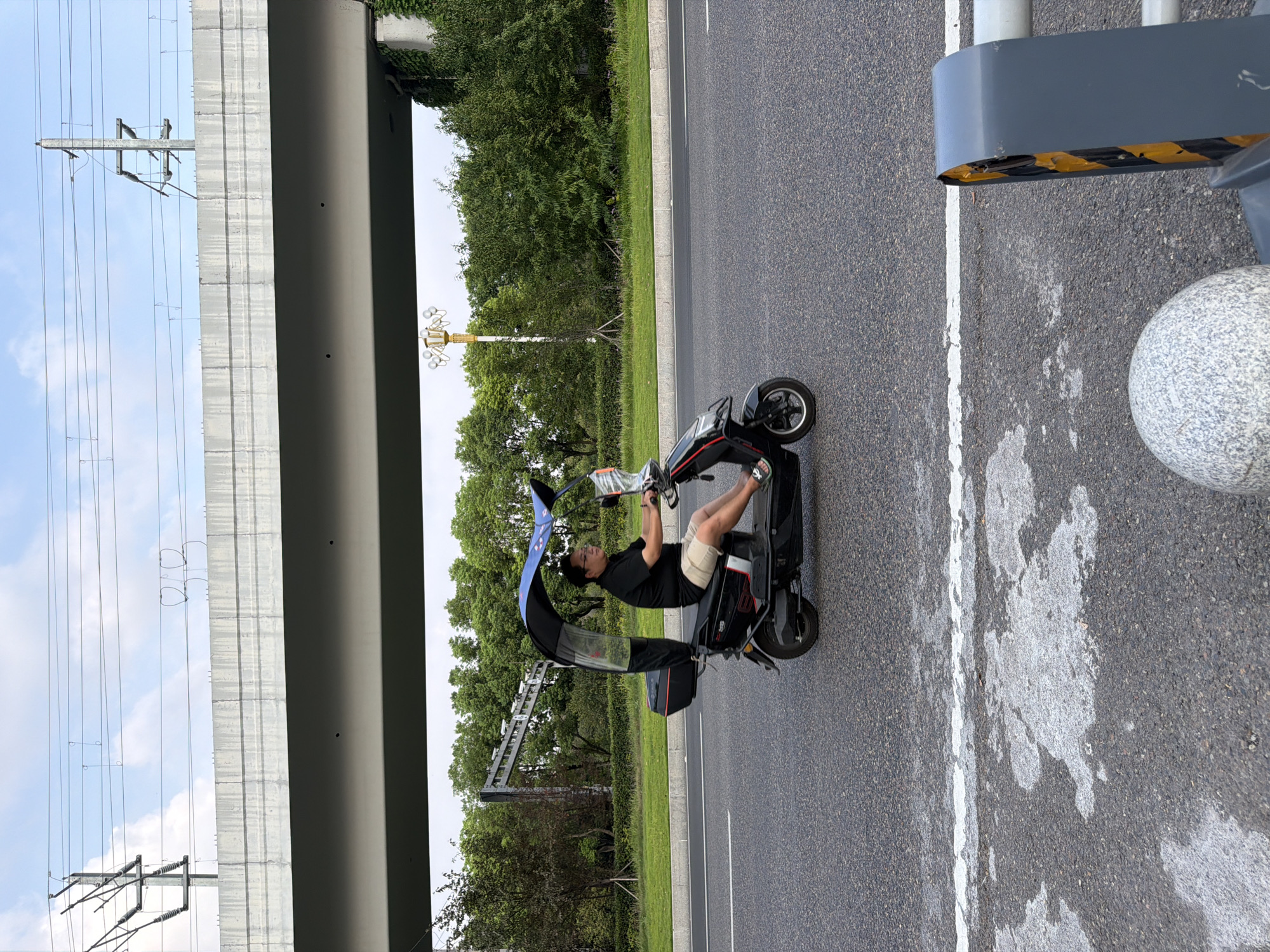 A man rides a black electric scooter with a blue canopy and clear side panels down a paved road in Jiangjin, China. He is wearing a black polo shirt and tan shorts. The scooter has a red taillight and a black storage box. The road is bordered by a grassy median with trees in the background. Above the trees is a concrete overpass with electrical wires running overhead. The sky is partly cloudy.