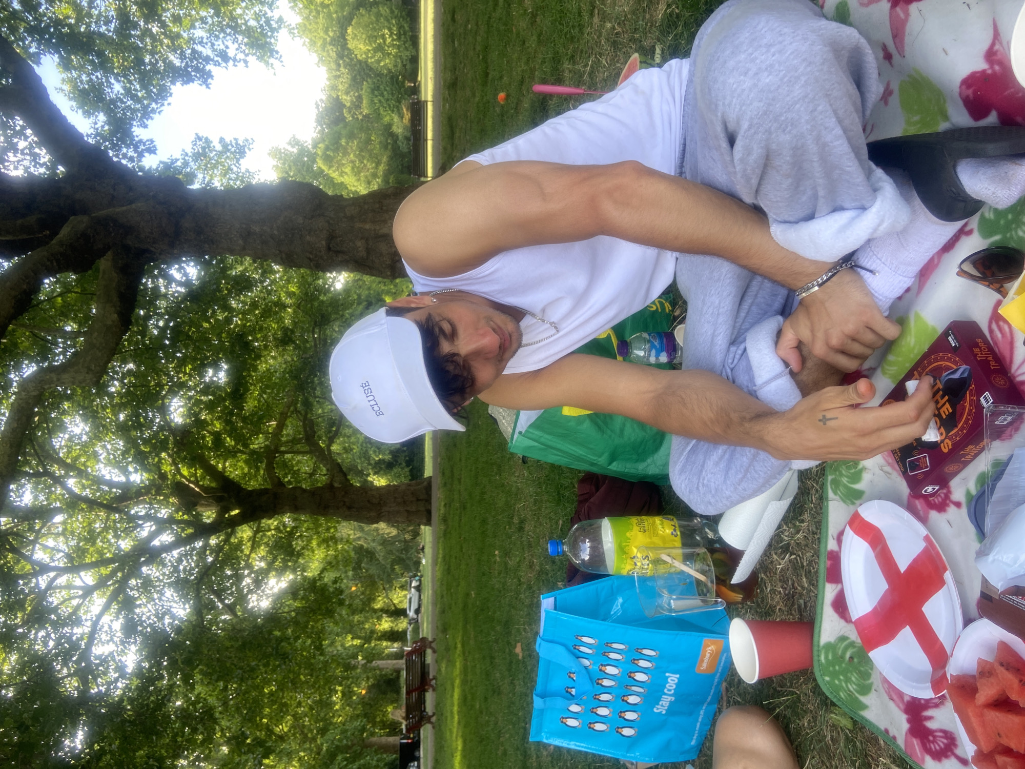 A young man wearing a white cap, white tank top, and grey sweatpants sits on a picnic blanket in a park, facing away from the camera. He is looking down and appears to be holding something in his hands. Scattered around him on the blanket are various items, including a blue shopping bag with penguins on it, a bottle of soda, a pitcher of a dark liquid, a red cup, sliced watermelon, a small box with the word "THE" visible, and a plate with a red and white cross design. To the right of the man, a pink object, possibly a toy or a prop, is visible. In the background, there are trees, green grass, and park benches, suggesting an outdoor setting. The presence of the England flag design on the plate and the context of a park picnic in London, United Kingdom, are implied by the elements in the image.