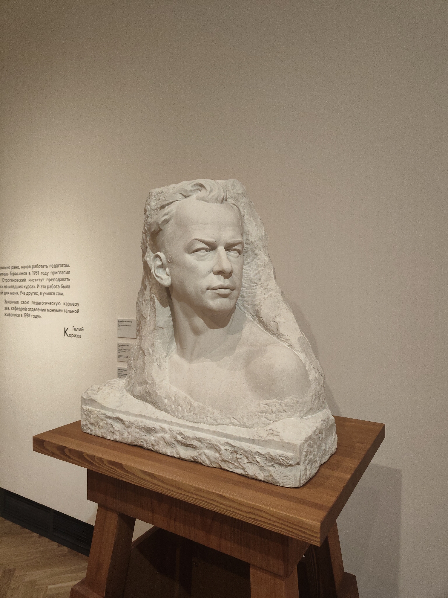 A white, stone bust of a man is displayed on a light-brown, wooden pedestal in front of a light-beige wall. The man has a light complexion, short, combed-back hair, a strong brow line, and a serious facial expression. The bust appears to be carved from a single, roughly-hewn block of stone. To the left of the bust on the wall, there is a text in a foreign language. The floor is dark and out of focus. The location is Unknown, Unknown.