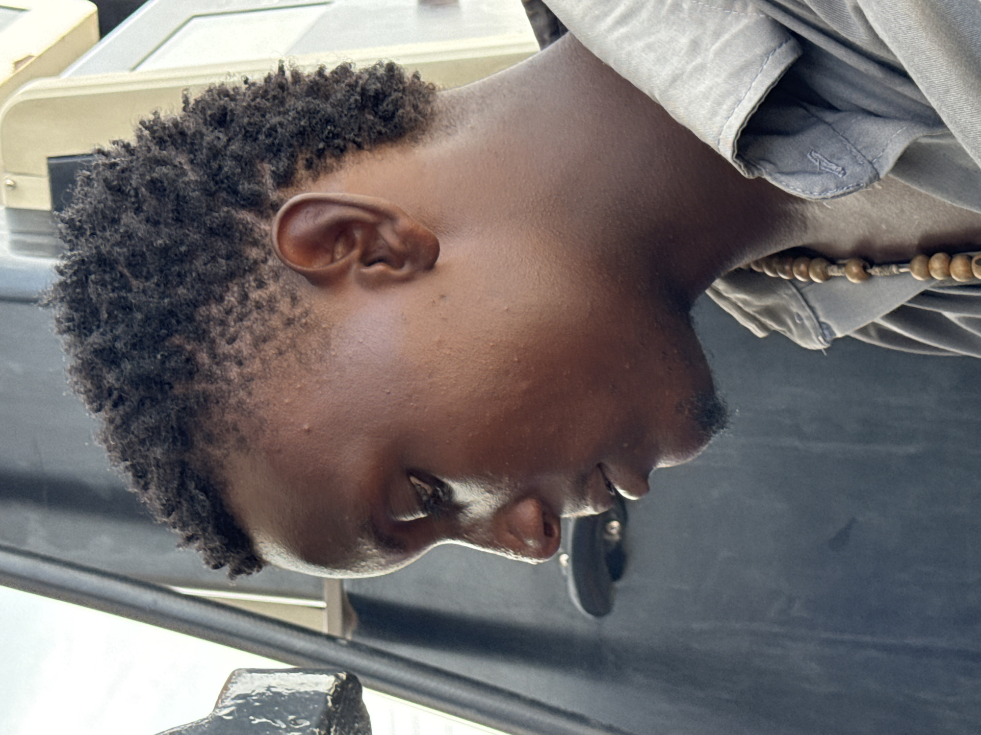 The image shows a close-up, rotated view of a dark-skinned man's head and upper chest. He has short, dark curly hair and is wearing a light-colored shirt with a beaded necklace. The background is blurred, with parts of a gray surface and a metallic object visible. Based on the information, the image is from Ado, Nigeria, but without further contextual information, the specific location within the city cannot be determined.