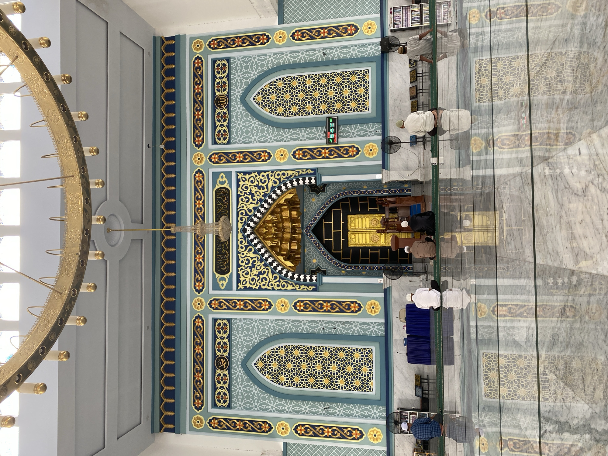 This media file captures the interior of a mosque in Lhokseumawe, Indonesia. The scene shows several individuals engaged in prayer. The architecture features ornate Islamic geometric patterns in shades of teal, blue, and gold adorning the walls. A large, elaborate golden chandelier hangs from the ceiling on the left side of the frame. The floor is a polished marble with reflective qualities, mirroring the intricate designs of the walls and the people praying.
In the foreground, the curved golden structure of the chandelier is prominent, with multiple light fixtures. The wall behind it is decorated with a repeating pattern of blue borders and gold floral motifs, interspersed with Arabic calligraphy within circular frames. The central section of the wall is dominated by a large arched niche with a golden geometric screen, and below it, a smaller rectangular niche with Arabic text in a darker frame.
Further into the mosque, several men are seen in prayer. They are dressed in traditional attire, including white thobes and skullcaps. Some are kneeling or prostrating on prayer mats, while others are standing. Two electric fans are visible, suggesting a warm climate. Small blue tables are set up in front of some of the prayer spaces.
On the right side of the image, a bookshelf filled with religious texts is partially visible. The overall atmosphere appears peaceful and devout. The lighting is bright, indicating it is likely daytime. The polished marble floor reflects the surroundings, adding depth and a sense of grandeur to the space. The image is oriented vertically.