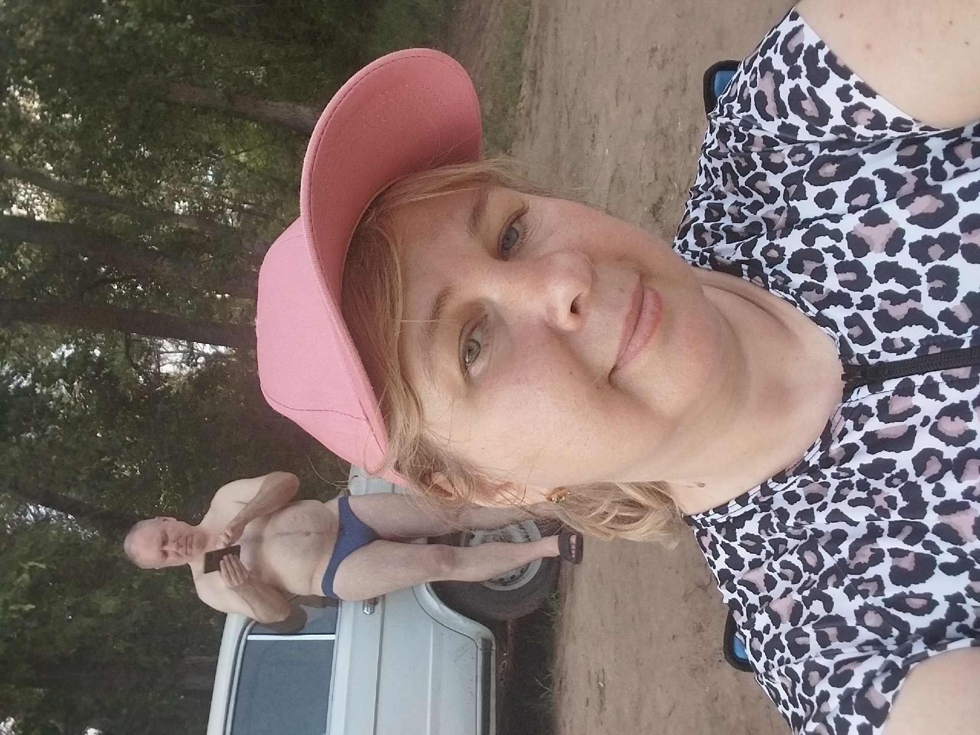 In Chkalov, Russia, a woman in a pink baseball cap and a leopard-print top is taking a selfie. Behind her, a man wearing blue briefs and holding a smartphone stands by a white car, with trees and foliage forming the background. The ground appears to be dirt.