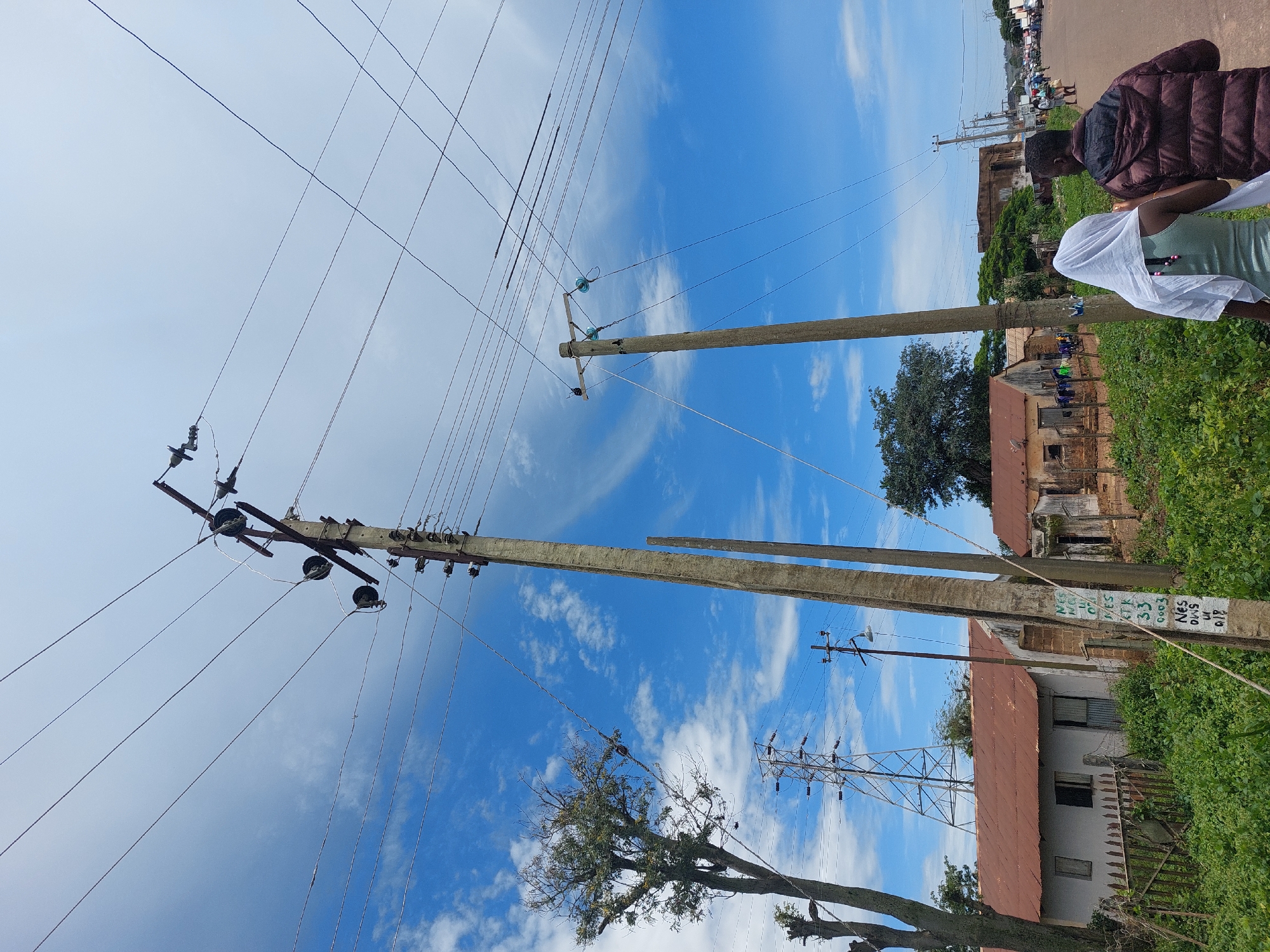 A street-level, eye-level, outdoor, slightly low-angle, full shot captures an overcast sky and a clear blue sky with scattered white clouds over a neighborhood in Barakin, Nigeria. Two individuals, a woman in a white headscarf and a man in a maroon hooded jacket, walk away from the viewer on the right side of a paved road. In the foreground, a tall, weathered wooden utility pole with multiple crossbars and insulators is situated on the left side of the road, next to a dense growth of green vegetation. A second, similar pole stands a bit further back on the right. Power lines crisscross the sky from the poles. To the left of the foreground pole, a tall, sparse tree with exposed branches and a few green leaves stands beside a metal transmission tower. Behind the vegetation, several low-slung buildings with rusted, reddish-brown metal roofs are visible. One building has a white facade and a weathered wooden fence. Another building appears to have an open doorway and clothes hanging on a line. In the background, more buildings and trees line the road, and several vehicles and pedestrians can be seen further down the street. The overall scene depicts a residential area with visible electrical infrastructure.