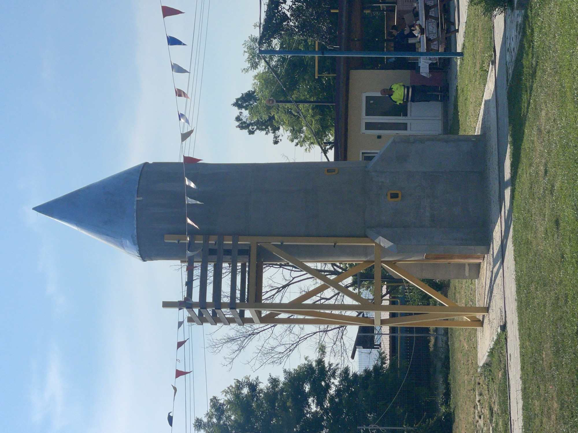 The image depicts an outdoor scene in the city of Zîrnești, Moldova, featuring a conical structure resembling a rocket, possibly a playground element or a decorative object, with a wooden scaffolding attached to its side. Small triangular flags in red, white, and blue are strung across wires above the structure, suggesting a festive or event-related atmosphere. In the background, a light-colored building with white doors and windows is visible, with a man in a bright yellow and green vest standing in front of it. Several picnic tables with checkered tablecloths are set up in front of the building, with some people seated at them. Trees and green grass form the surrounding environment, with a clear blue sky overhead.