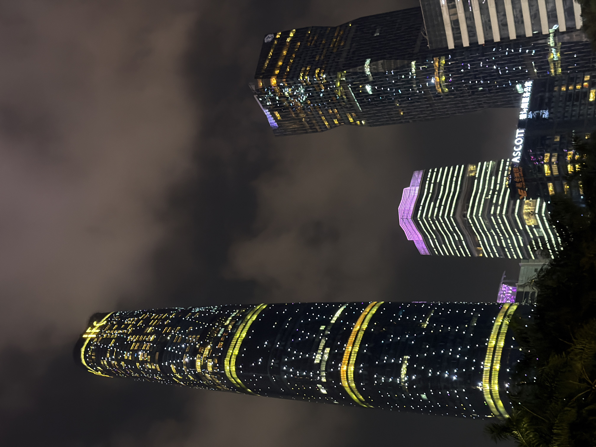 The image shows three illuminated skyscrapers in Tianhe District, China, at night. The Canton Tower, a cylindrical building with yellow bands, is at the bottom. Above it is a building with vertical white lines and purple accents on the top. The third skyscraper, at the top of the frame, has a darker facade with numerous yellow and white lights and the name "ASCOTT" on its side. Dark, cloudy sky surrounds the buildings, and some trees are visible on the right side.