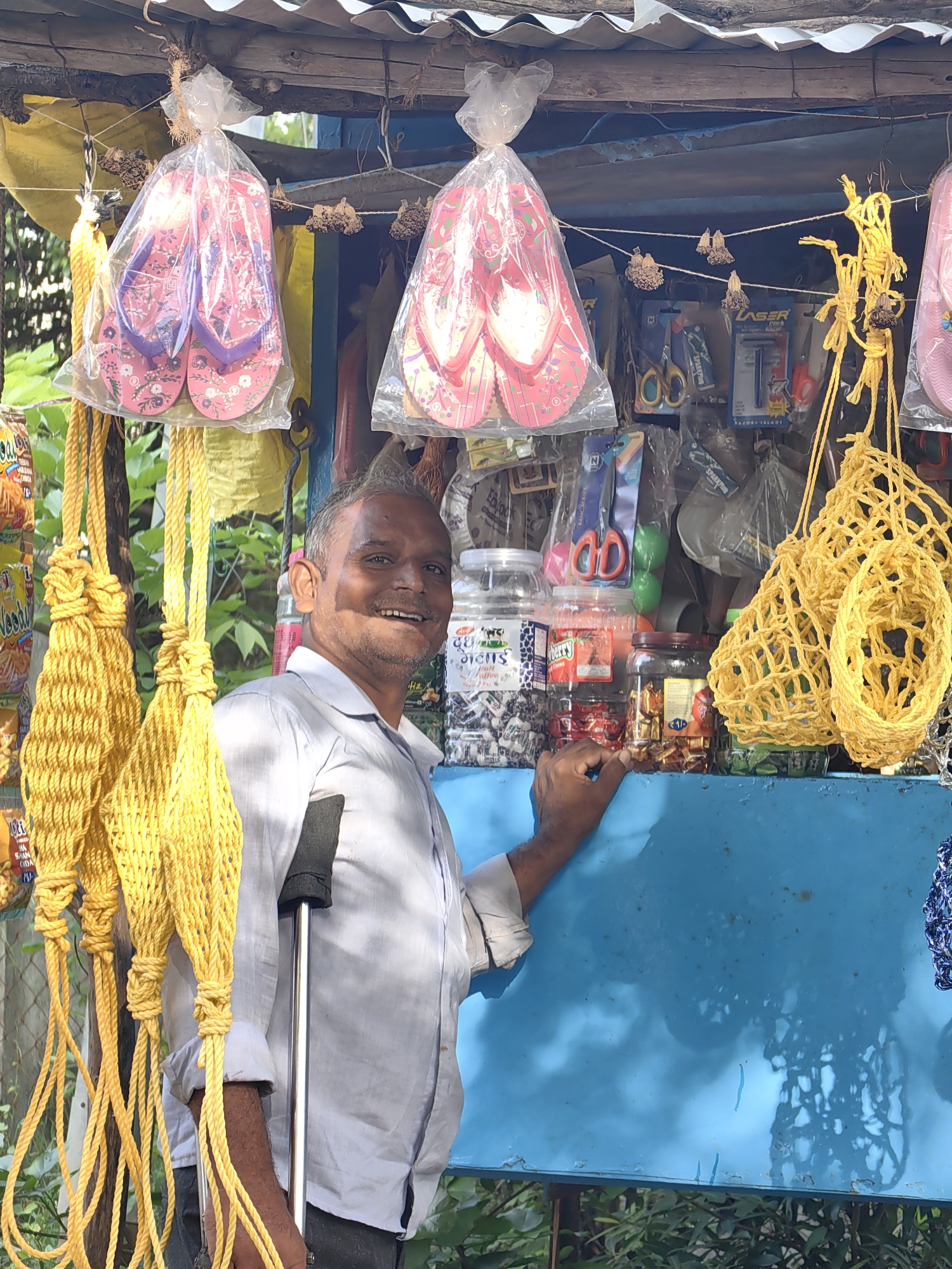 In Mhaiswadi, India, a man stands behind a stall displaying various items. He is leaning on a cane and smiling, with his right hand resting on the blue surface of the stall. The stall itself is covered by a corrugated metal roof. Hanging from the stall are several pairs of pink flip-flops in plastic packaging, along with yellow braided ropes and woven yellow nets. Shelves behind the man are stocked with jars of what appear to be snacks and small packaged goods, including scissors and razors. The background shows greenery and other stalls.