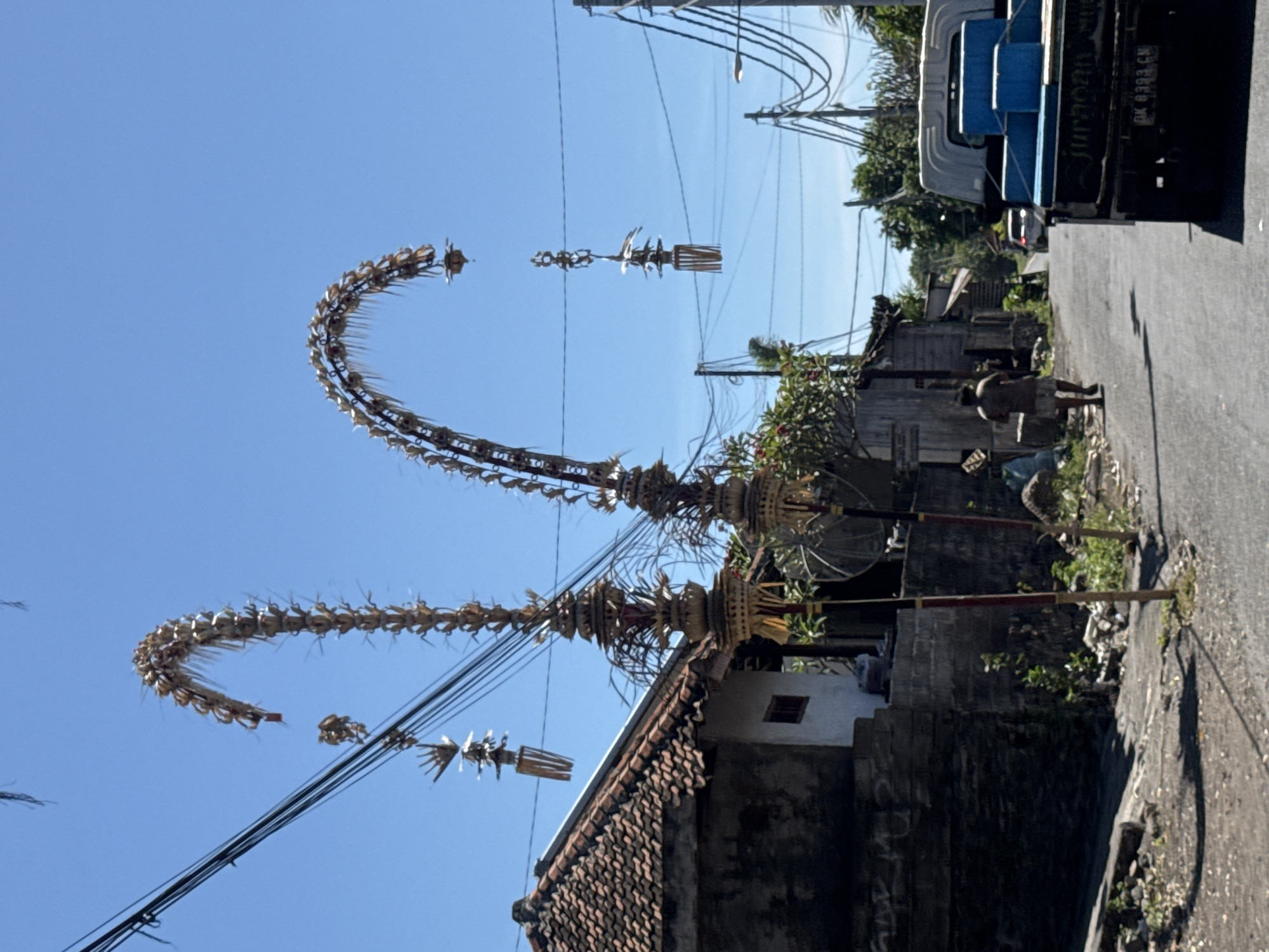 The image shows two Penjors erected on the side of a road in Kubu, Indonesia. These are tall, curved bamboo poles decorated with intricate woven palm leaves and offerings. Power lines stretch across the sky above. A tiled roof of a building is visible below one of the Penjors. A person walks along the side of the road, and a portion of a truck is seen parked on the right side of the frame.
