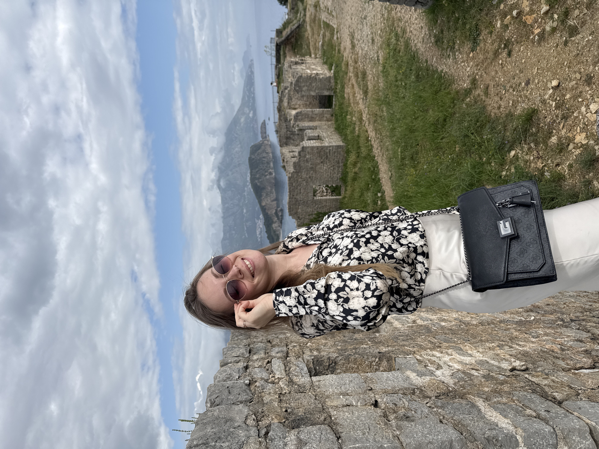 A young woman stands on a stone wall, smiling and wearing sunglasses, a floral blouse, and white pants. She has a black purse on her shoulder. Behind her is a view of the sea, islands, mountains, and cloudy sky. A stone building is visible to the right of her. The location is the Citadel in Budva, Montenegro.