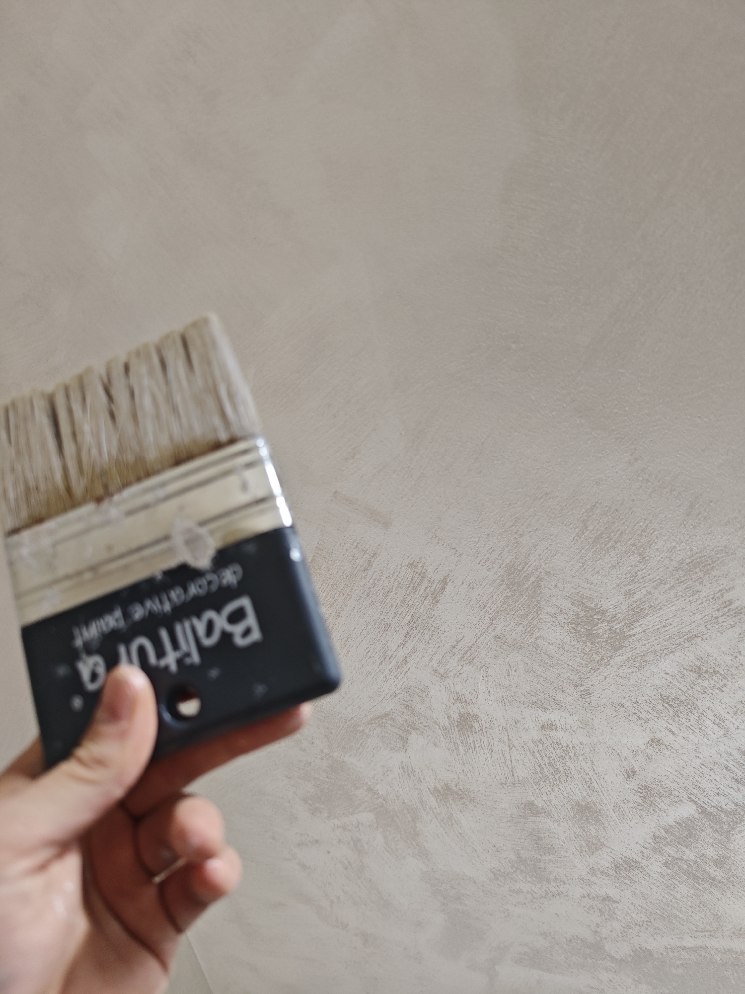 A close-up shot captures a hand holding a paintbrush against a textured wall. The hand, with light skin, grasps the dark blue-grey handle of the brush. Visible text on the handle, inverted, reads "Baltura" and "decorative paint" beneath it. The brush has light-colored bristles that appear to be used, possibly with dried paint matching the wall's hue. The wall itself is painted in a light, neutral tone, likely beige or light grey, and features a distinct decorative finish with prominent, sweeping brush strokes that create a textured, non-uniform surface. The image depicts the ongoing activity of applying decorative paint to an interior wall. This scene is set in Sofia, Bulgaria.