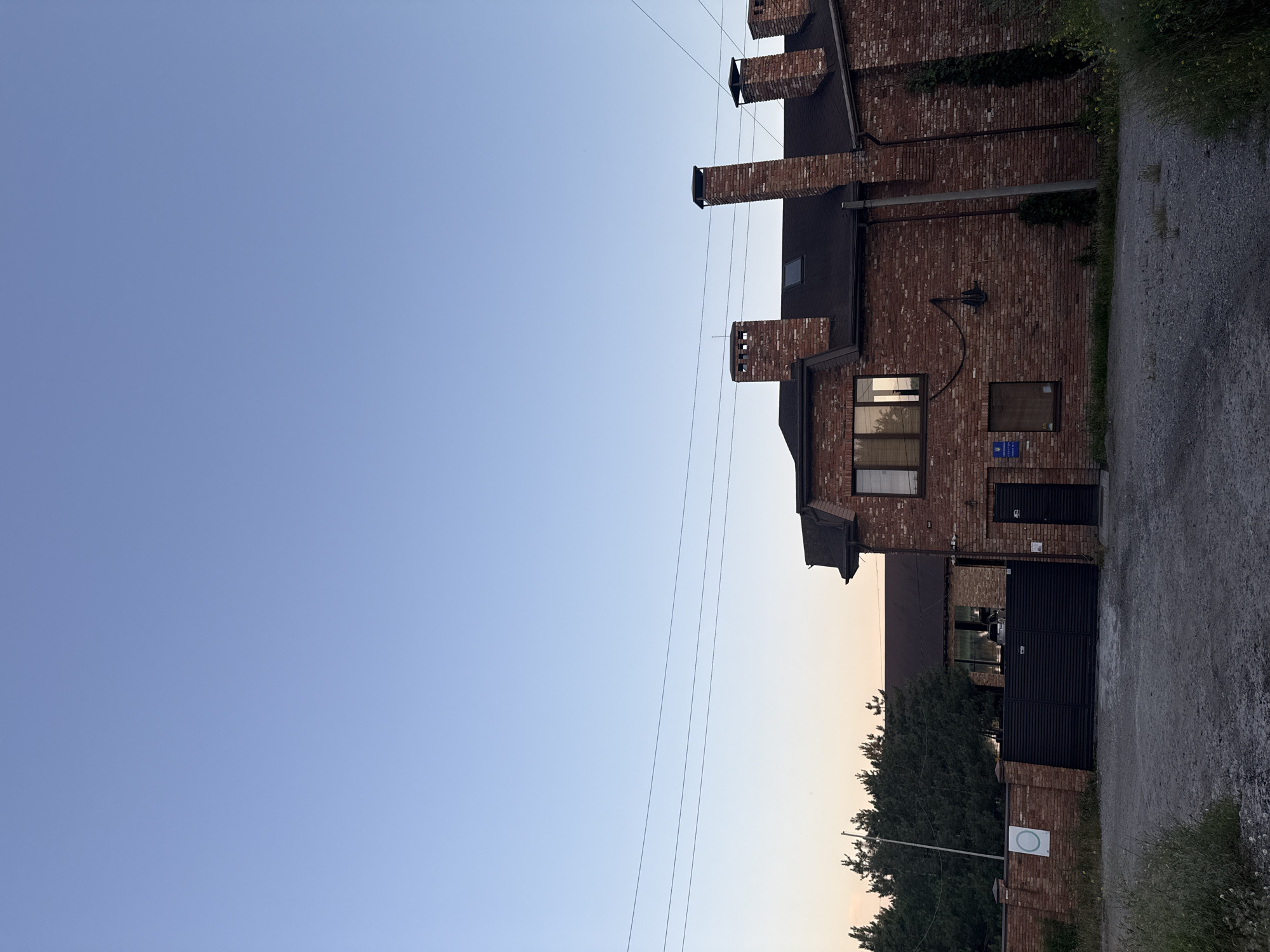 The image shows a large, two-story brick building with multiple chimneys, located in Obukhivka, Ukraine. The building has a dark roof and several windows. In front of the building is a black metal gate and a brick fence. Behind the fence, there are trees. The sky is clear and blue, with some power lines visible. The scene appears to be a private residence or a business establishment. There are no people visible in the image. The ground in front of the building is covered with gravel.