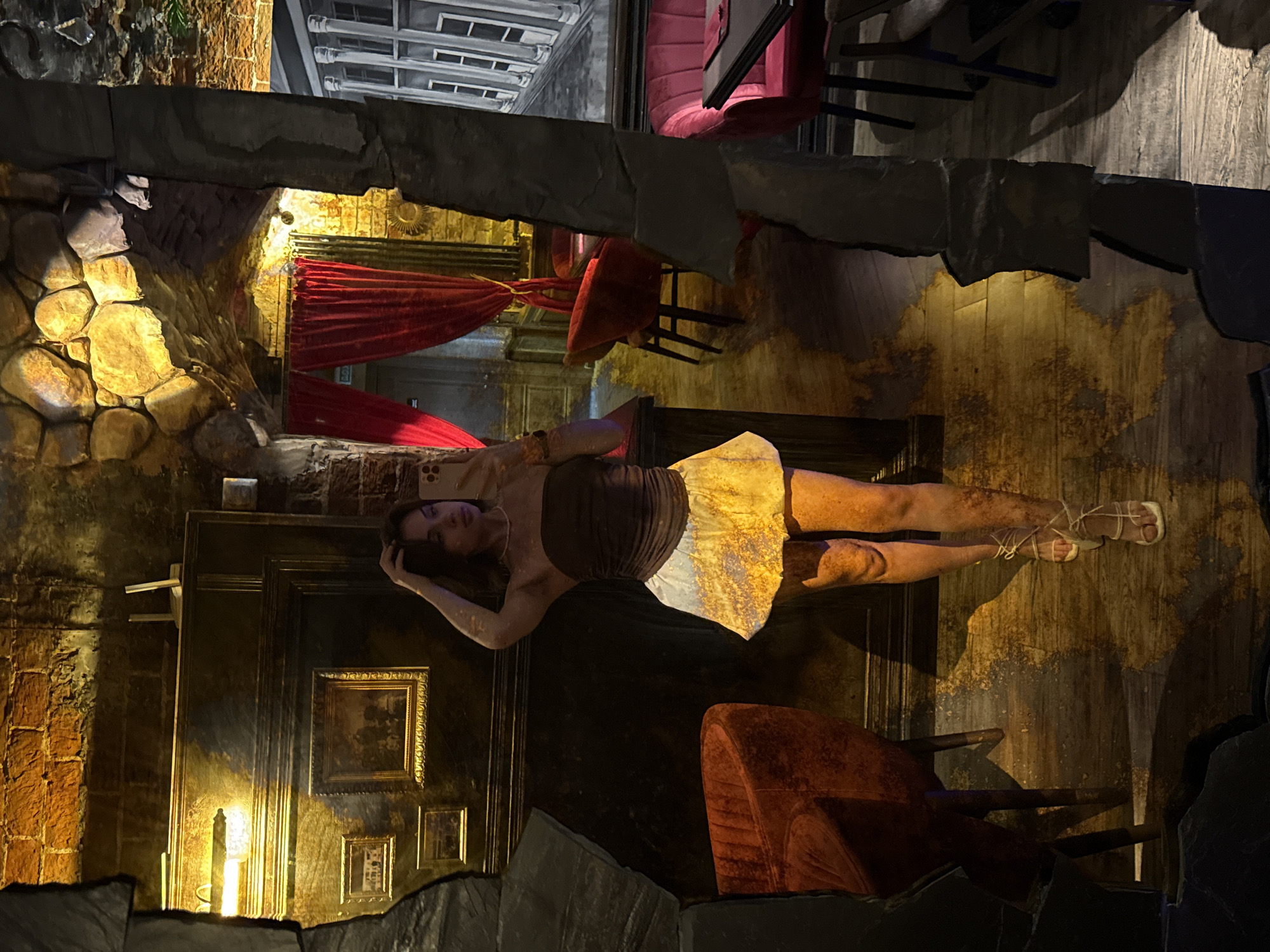 The image shows a woman reclining on a surface that appears to be a table or counter, oriented vertically in the frame. She is wearing a dark top and a light-colored skirt. The surrounding environment has a theatrical or stage-like quality, with stone walls on the left and a backdrop suggesting an interior room with a red curtain and pictures on the wall. The floor is a wooden plank surface, and there are chairs in the scene. The lighting is low and dramatic, with a warm color cast. It is difficult to ascertain the specific location in Sarapul, Russia, as it appears to be a constructed or theatrical setting.