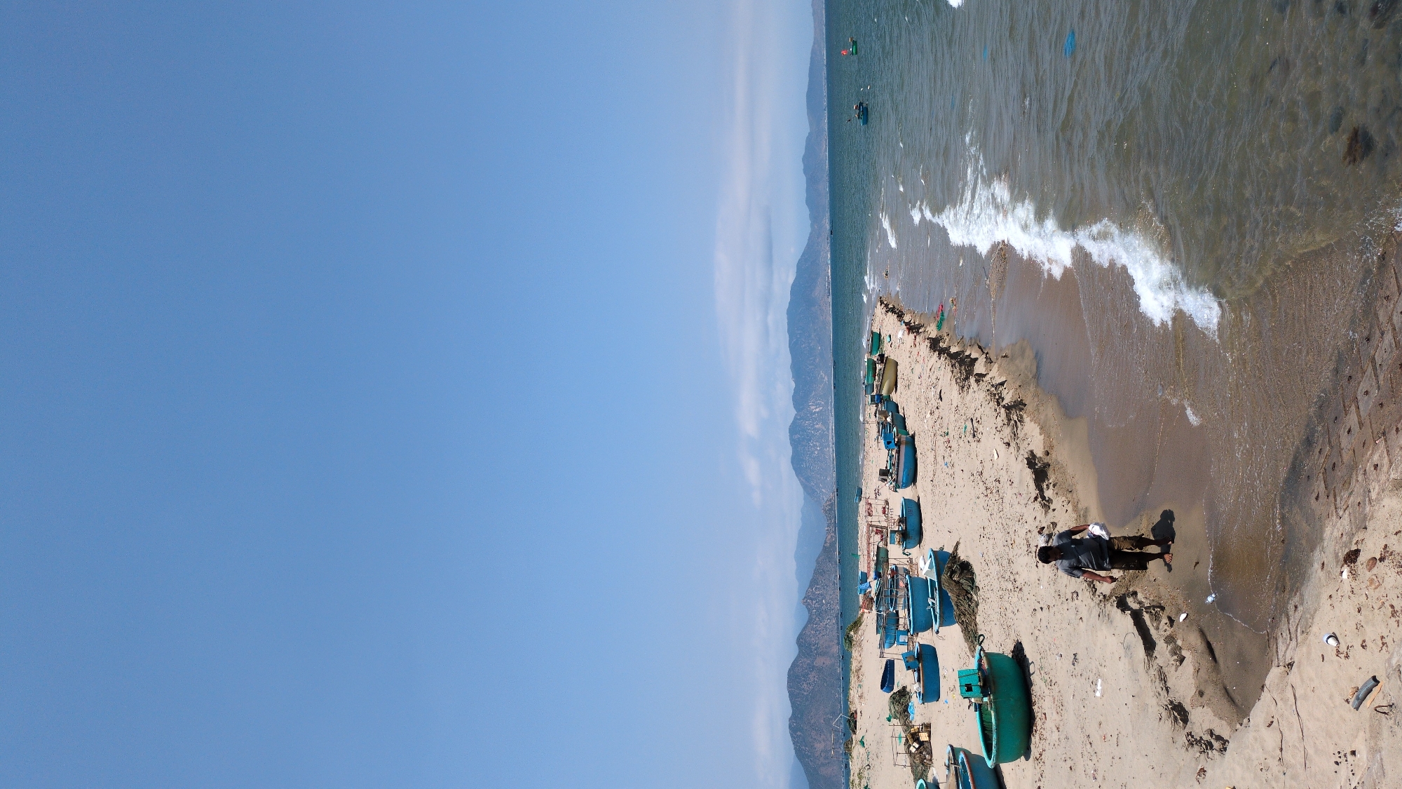 A wide-angle aerial shot shows a coastline with various details. The sky is clear blue. The shoreline has a mix of sand and water, with waves crashing onto the shore. The water is a murky green-brown color. A single person is visible standing near the water's edge, casting a shadow. Numerous blue and green boats are scattered along the sand. In the background, there is a mountainous region with light cloud cover near the peaks. Debris and objects litter the beach. The location is Phan Rang – Tháp Chàm, Vietnam.