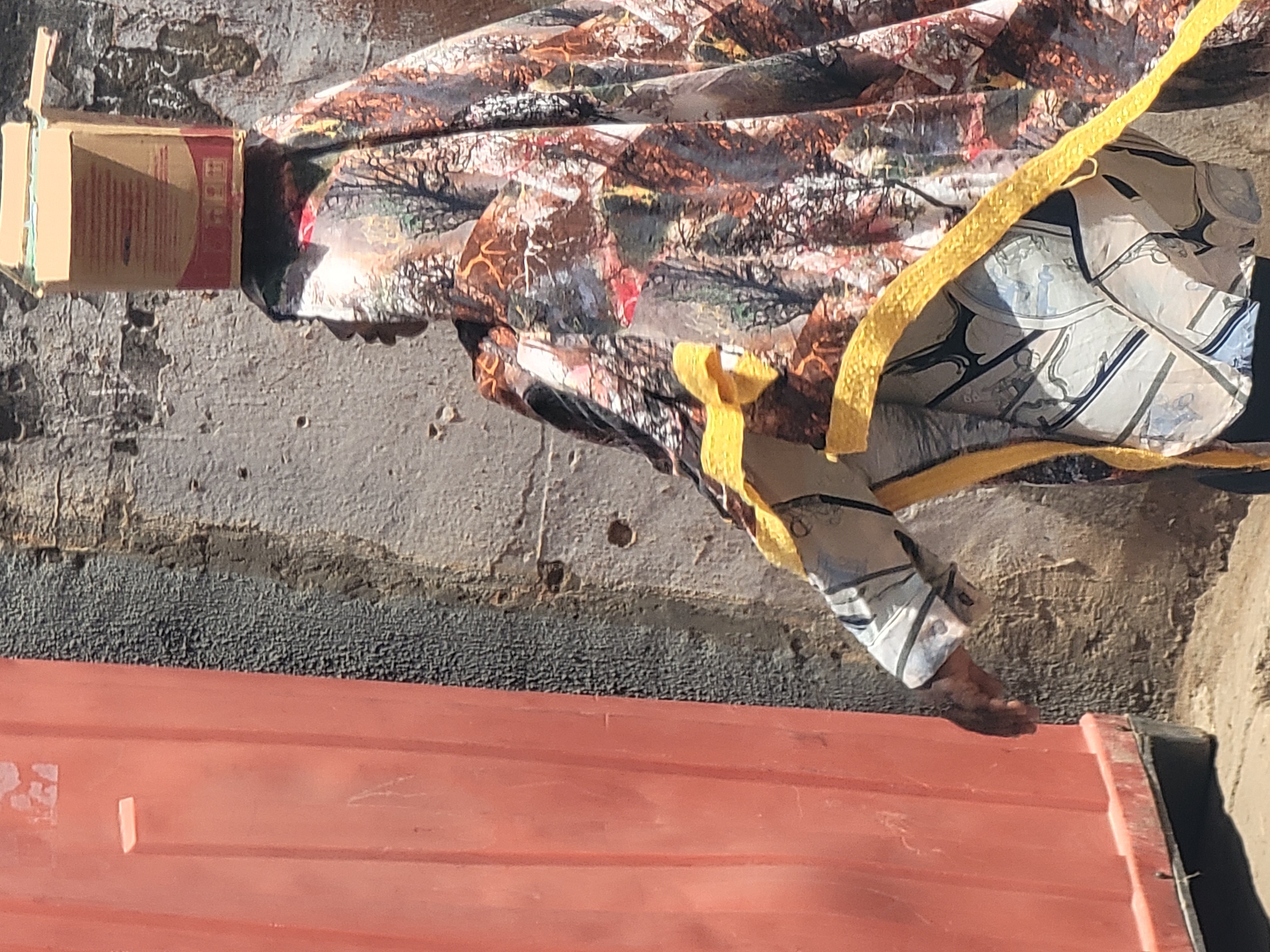 A human figure stands next to a red corrugated structure, possibly a shipping container, in an outdoor environment. The figure wears a patterned garment and has a yellow rope or strap wrapped around its body. A small cardboard box is positioned near the figure's head. The background includes a wall or surface with visible texture and discoloration. The location is presumed to be Maiburiji, Nigeria.
