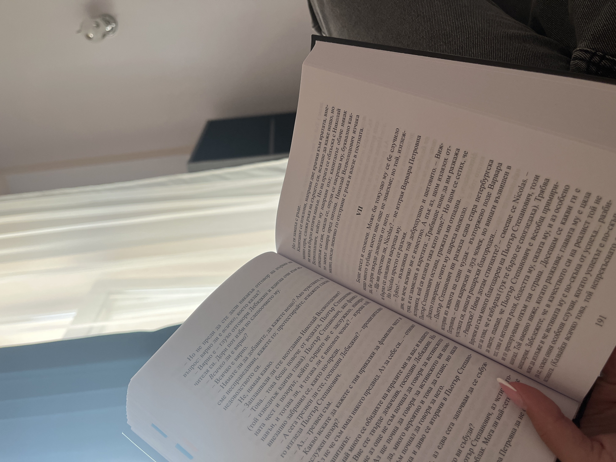 In an indoor environment, a book with pages partially filled with Bulgarian text is held open by a person's hand. The book's pages feature dense paragraphs of text and some line breaks. The right page displays a chapter heading, "VII", in a larger font. The background shows a portion of a room with light-colored curtains and a wall with a small light fixture. The overall impression is of someone reading or about to read. There are no discernible Plovdiv landmarks or specific city indicators.