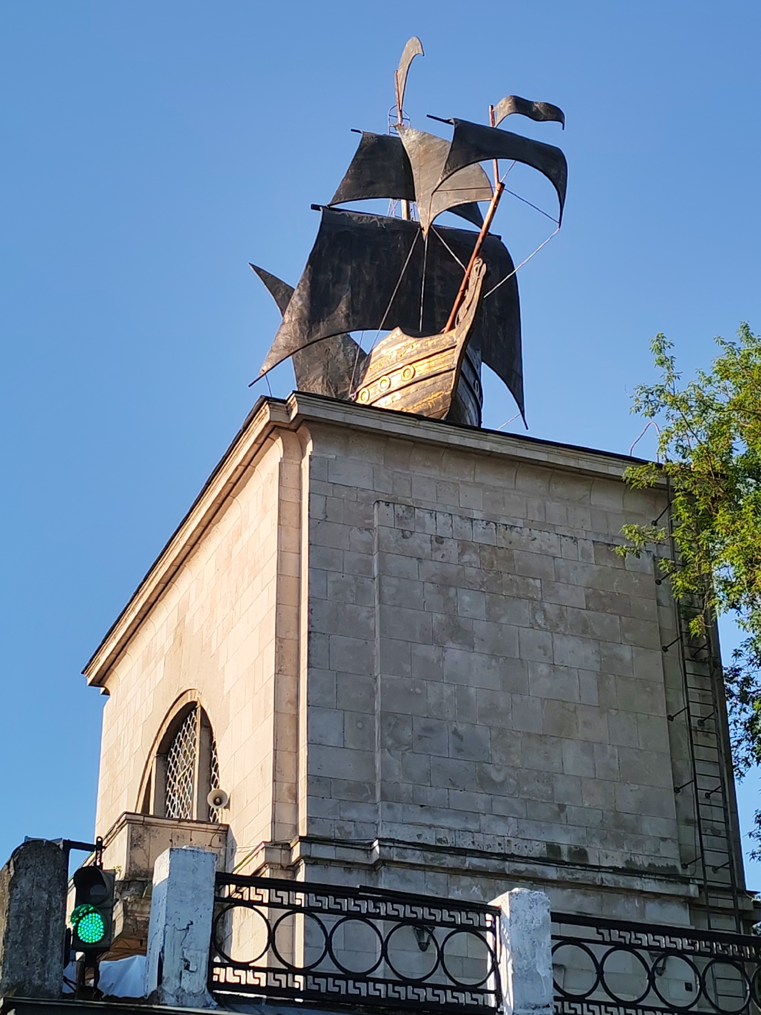 The image shows the Yakhroma water tower in Yakhroma, Russia. The tower is a tall, rectangular structure built of light-colored stone blocks. It has a decorative metal ship sculpture on its roof. A green traffic light is visible at the bottom left corner. A black metal railing with circular designs runs along the base of the tower. A metal ladder is attached to the right side of the tower, partially obscured by a tree branch. The sky is clear and blue. A window with a decorative grill can be seen on the side of the tower. A loudspeaker is mounted near the window.