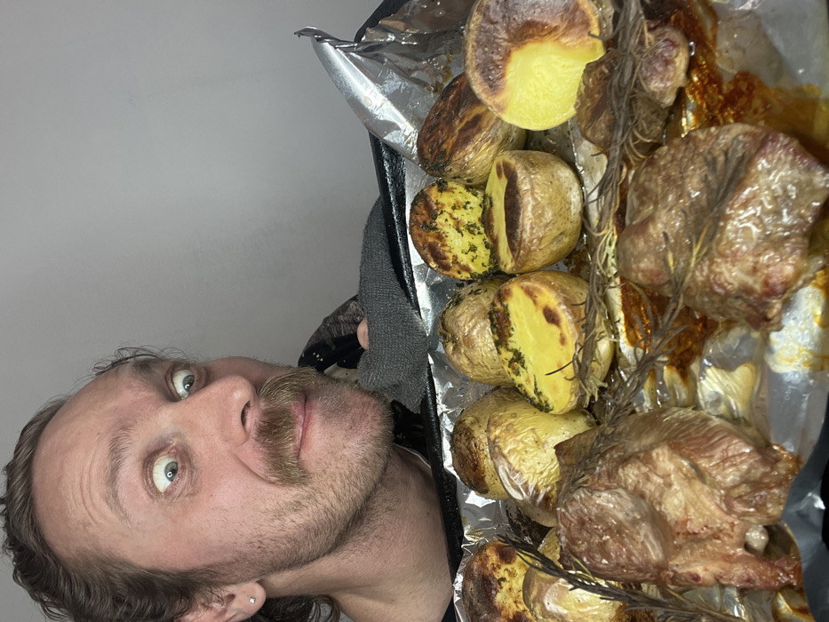 A male individual with light brown hair and a prominent mustache stares intently at a baking tray filled with recently cooked food. His eyes are wide open, and his expression suggests anticipation. He wears a dark garment, and a small silver stud earring is visible in his left ear. His left hand, covered by a gray oven mitt or glove, grips the edge of the black baking tray, which is lined with crumpled aluminum foil. The foil shows residue from rendered fat and spices.
The baking tray contains an assortment of roasted items. Numerous small, whole, and halved potatoes are visible, some with browned skins and others exposing their yellow interiors, often sprinkled with green herbs like rosemary or parsley. Several pieces of roasted meat, appearing to be lamb or beef, with browned exteriors and a reddish-brown hue, are also present, interspersed with fresh rosemary sprigs. The food is coated in visible oils and drippings. The background is a plain, light gray wall, indicative of an indoor setting. There are no visual cues within the image to determine the specific location in Saint Petersburg, Russia.