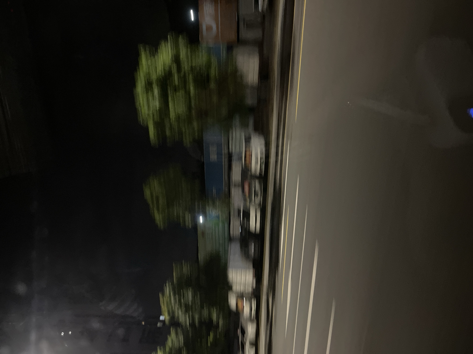 A nighttime, motion-blurred photograph captures a street scene in Panyu District, China. In the foreground, a paved road with white and yellow lane markings is visible. Beyond the road, a row of parked vehicles, including white trucks and cars, are blurred due to the motion of the camera. Trees with green foliage are also visible, heavily blurred, lining the street. In the background, large, light-colored buildings are present, with some signs or logos visible. The sky is dark, indicating it is nighttime. The overall impression is of a street in an urban area at night, taken from a moving vehicle.