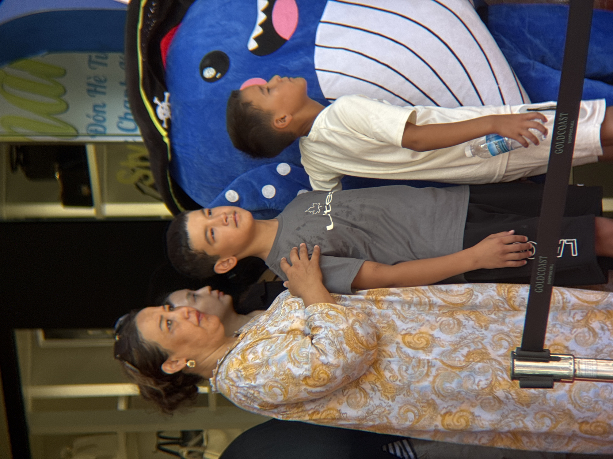 A group of four people, including two young boys and two women, are posing for a photo in front of a large blue whale mascot in what appears to be a commercial setting. The woman on the left is wearing a patterned dress and earrings, while the other woman is partially obscured. The two boys are wearing t-shirts, and one is holding a bottle of water. The blue whale mascot is dressed in a pirate hat with a skull and crossbones. In the background, there are shelves with merchandise, and a sign with Vietnamese text is visible. A stanchion with a black ribbon and the words "GOLDCOAST SHOPPING MALL" is in the foreground. The scene suggests a promotional event or a shopping mall attraction in Nha Trang, Vietnam.