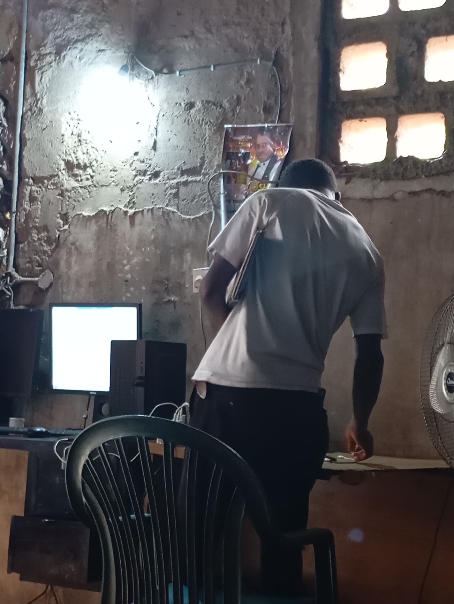 The image depicts an indoor scene, possibly in Shendam, Nigeria, characterized by a rough, textured concrete wall and a grimy window with multiple square panes. A single bare lightbulb hangs from the ceiling, casting a bright, localized illumination. In the foreground, a person, viewed from the back, is wearing a light-colored t-shirt and dark trousers. This individual appears to be leaning over a table or desk, holding what might be a laptop or folder.
Several computer monitors and computer towers are visible on a desk in the mid-ground, with one monitor displaying a bright white screen. A framed picture of a man in formal attire is mounted on the wall behind the person. To the right of the frame, a rotating fan is partially visible. A dark plastic chair is in the immediate foreground, partially obstructing the view of the desk and the person. The overall environment suggests a functional but somewhat unkempt space, likely used for computer-related activities.