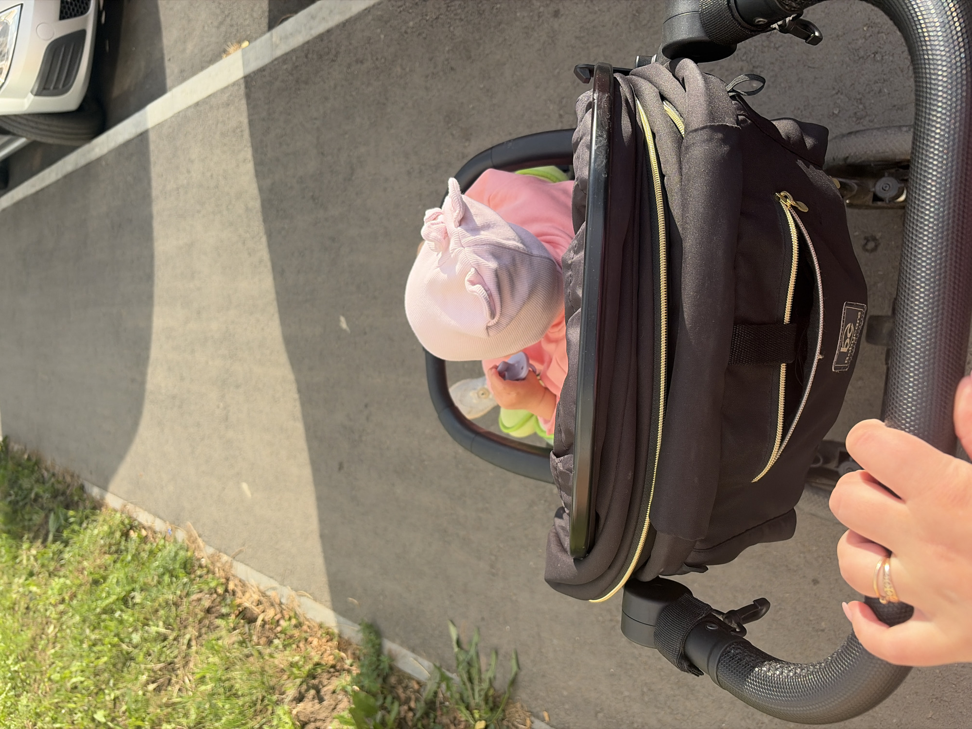 A first-person perspective shot shows a person's hand with a ring on their finger gripping the handle of a black stroller. The stroller contains a baby wearing a pink hat and pink clothing, holding a purple pacifier. The stroller is on an asphalt path next to a grassy area. A silver car is parked on the right side of the frame, with its front bumper and wheel visible. The scene appears to be a sunny day, casting shadows on the path. The location is identified as Izhevsk, Russia.