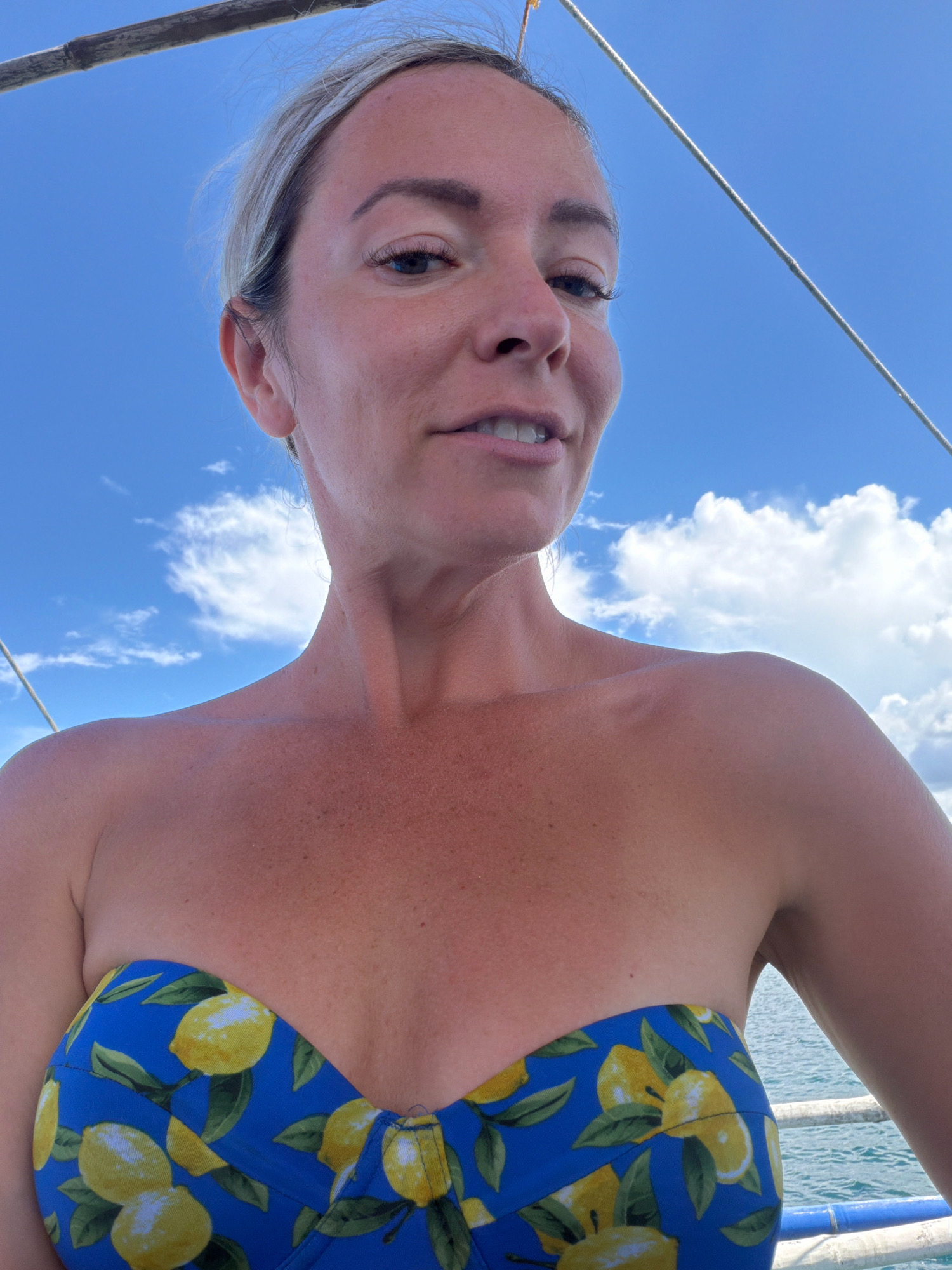 A woman is captured in a selfie-style shot, her face partially visible from a low-angle perspective. She appears to be on a boat, suggested by the rope visible at the top of the frame and the railing and water in the background. The sky is a vibrant blue with scattered white clouds, indicating a sunny day. The woman has blonde hair pulled back and is wearing a blue bikini top adorned with a yellow lemon print. The background water appears calm and tropical. The overall setting suggests a leisure activity in a coastal area, possibly a tourist destination known for boat trips or beaches. The woman's expression is slightly smirking, looking towards the camera. The lighting indicates it is daytime.