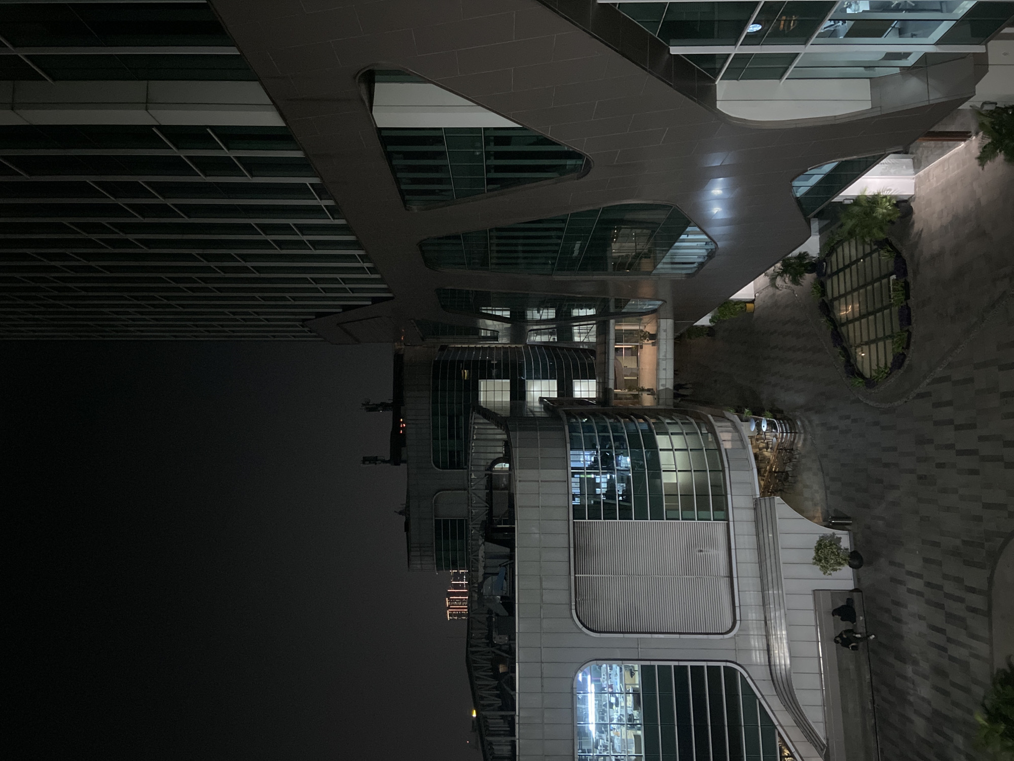 The image depicts an outdoor scene at night in what appears to be North Jakarta, Indonesia. A multi-story modern building with a glass facade dominates the view. A connecting structure with angular openings bridges to another part of the building. The area below is paved with patterned brick. A small, enclosed garden is visible. People are observed seated on a bench. In the background, city lights are visible against the dark sky.