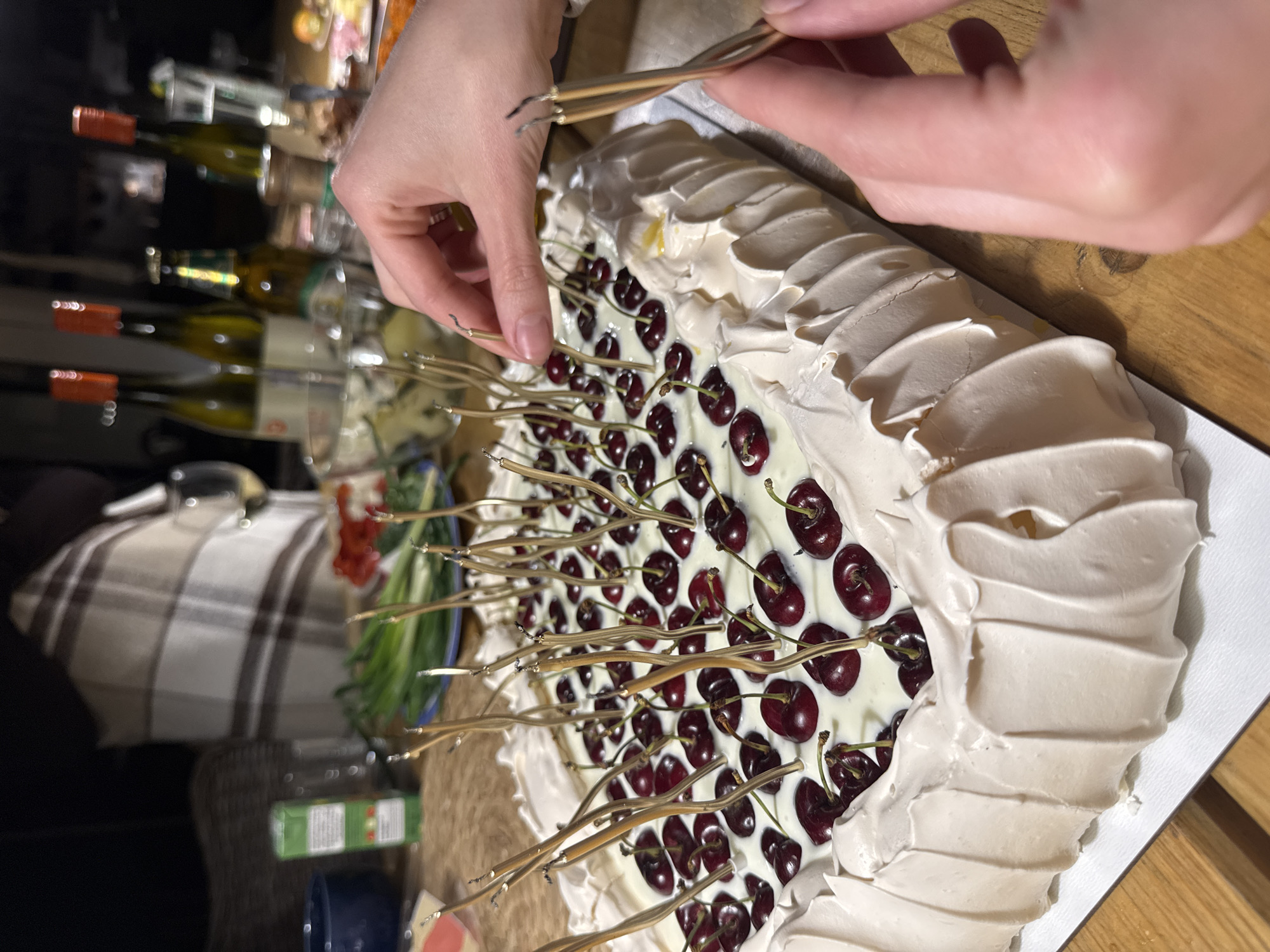 A rectangular-shaped cake is displayed, decorated with rows of dark red cherries, each with its stem still attached. A person is using tweezers to carefully place the cherries on top of the cake. The cake is adorned with a frosting in swirls. In the background, there are bottles of wine, a plaid tablecloth, and other items. The location cannot be verified.