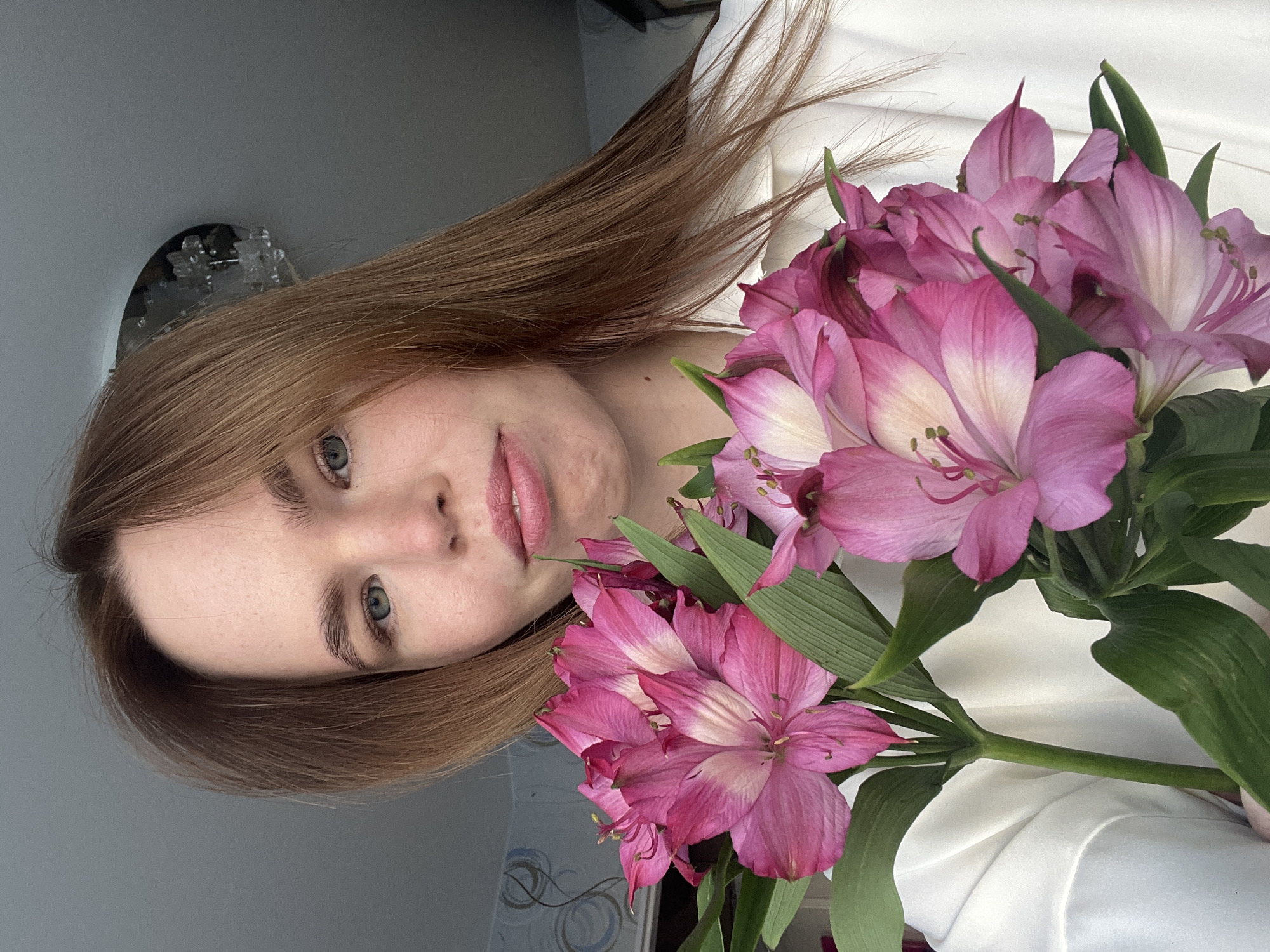 The image presents a close-up portrait of a young adult female situated indoors. The subject has long, light brown hair, blue-grey eyes, and fair skin with visible freckles, looking directly at the camera with a slight smile. She is wearing a white collared shirt. Held in front of her, partially obscuring her lower body, is a bouquet of pink and white Alstroemeria flowers, characterized by multiple blooms with vibrant pink petals that transition to lighter centers, accompanied by green stems and leaves. The background features a light-colored wall, with a section of light blue-grey patterned wallpaper visible on the left side and a plain light grey area on the right. A portion of a translucent, decorative ceiling light fixture is visible in the upper right quadrant of the image. The specific location within the city of Chaikovsky, Russia, cannot be determined from the visual evidence provided.