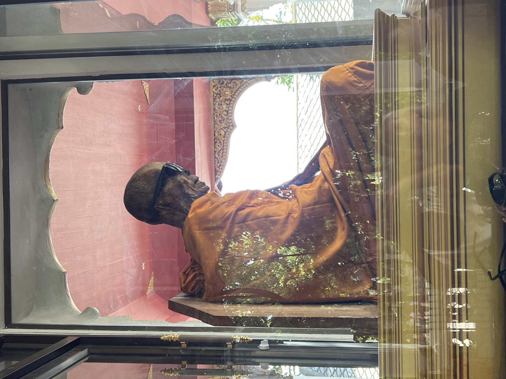 The image shows a mummified monk in a seated position, wearing orange robes and sunglasses, displayed inside a glass case. The monk is positioned on a wooden chair within the case, and behind him, there is a red decorative ceiling with intricate patterns. The background visible through the glass includes greenery from trees and a tiled roof structure, suggesting an outdoor or semi-outdoor setting. Reflections on the glass indicate the presence of other people and objects outside the case, including a detailed view of a golden pedestal on which the glass case rests. The overall scene appears to be within a temple or a similar religious or cultural site in Baan Hua Thanon, Thailand.