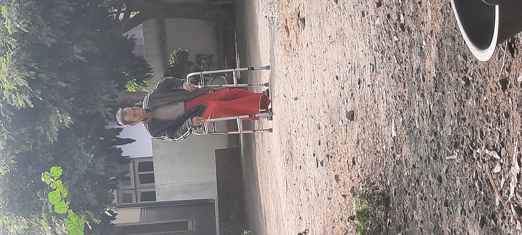 A man walks using a walker in an outdoor area. The man is wearing a black jacket with white stripes on the sleeves, a gray shirt, and red pants. He has short, dark hair and appears to be middle-aged or older. The walker is a silver, foldable frame with four legs. The ground is covered in dirt and small stones. In the background, there is a building with windows and a door, and a large tree with green leaves. The scene appears to be in a residential area. The specific location is the city of Selopuro, Indonesia.