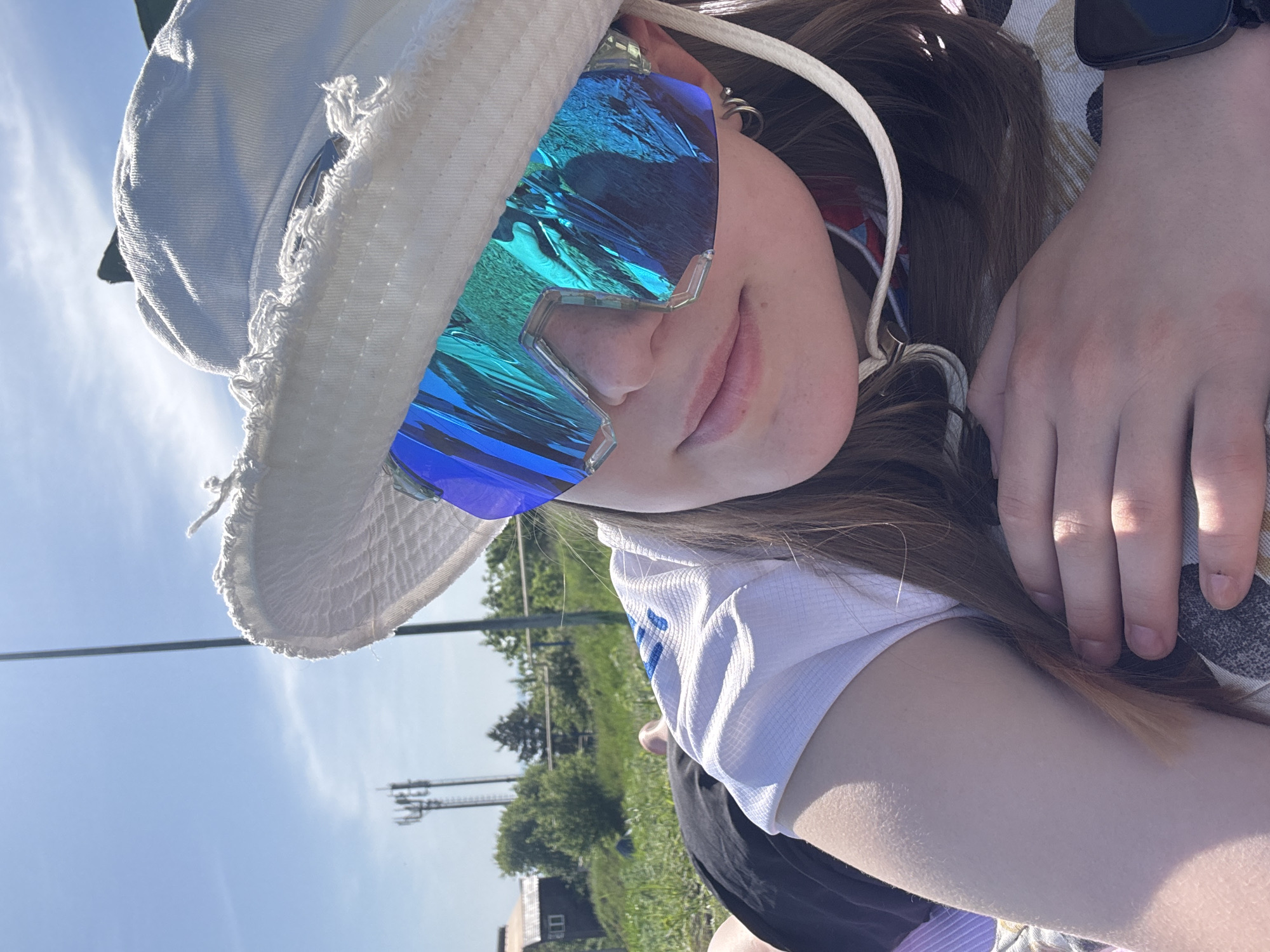 A person wearing a white bucket hat and blue and turquoise sunglasses poses for a photo. The subject is leaning slightly to the left. The background appears to be outdoors, featuring green grass, trees, and an infrastructure tower. The location is identified as Khvalynsk, Russia.