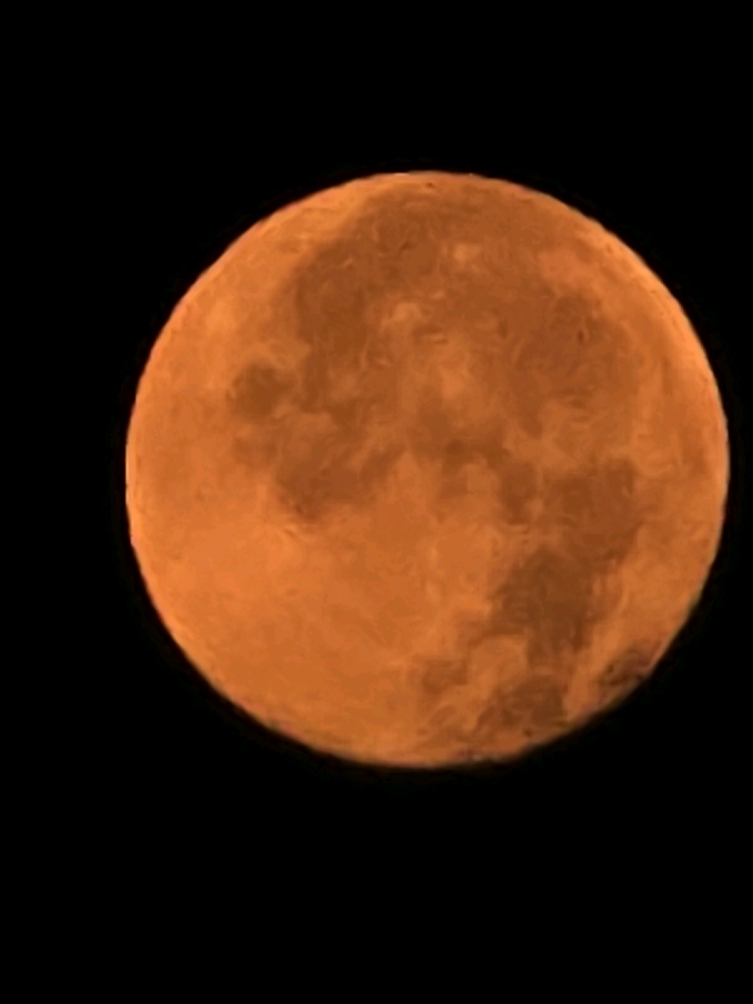 The image is a close-up, slightly blurry photograph of a large, orange celestial body against a black background. The celestial body appears to be the moon during a lunar eclipse or with atmospheric distortion, giving it a reddish-orange hue. Surface features, resembling craters and maria, are visible as darker and lighter patches across its disc. The composition focuses solely on this object, with no other elements present in the frame. The image does not depict any recognizable landmarks or scenery from Podgorica, Montenegro.