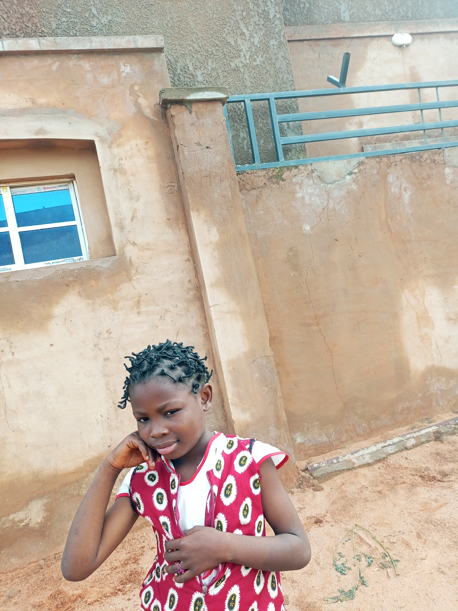 A young Black girl stands in front of a textured, beige building in Tempure, Nigeria. She has her hair styled in small braids and is wearing a white shirt with a red vest adorned with a white, circular pattern. Her left arm is bent, with her hand resting on her cheek as she looks towards the camera with a slight smile. A blue-tinted window with a white frame is visible on the left side of the building. A light blue metal railing runs horizontally along the upper part of the building's exterior. The ground is covered in sand.