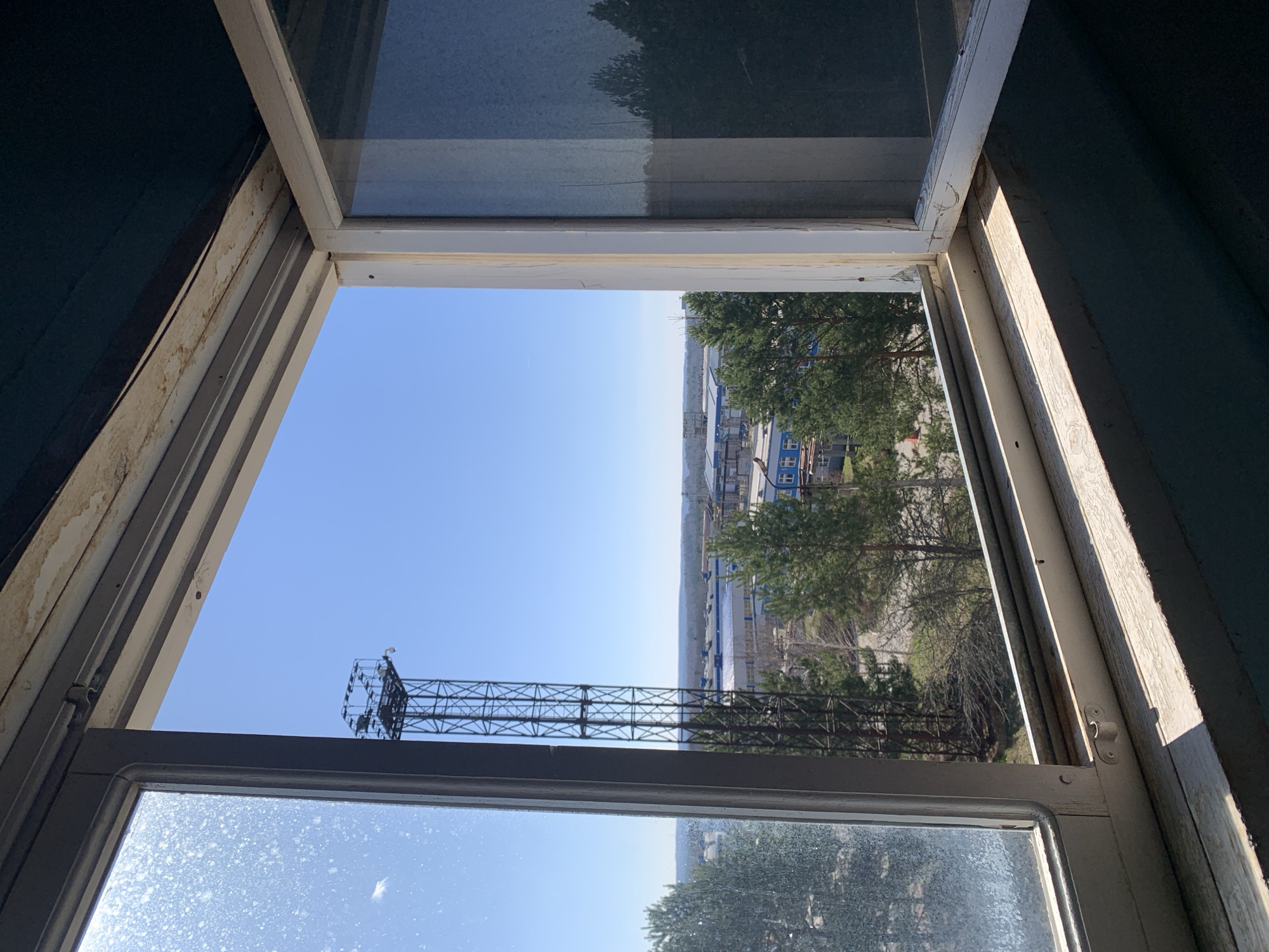 The image shows an open window with a view of a cityscape. In the foreground, the window frame is white and appears old, with visible wear and tear. The window is opened in a way that the top and bottom panes are pushed outwards, creating a trapezoidal opening to the outside.
The view through the window reveals a bright, clear sky above a landscape featuring buildings, trees, and a large metal structure that appears to be a communication tower.
Based on the known environment, the landscape appears to be an industrial one. The combination of the industrial facilities and the communication tower is consistent with the infrastructure often seen in a city like Chaikovsky, Russia.