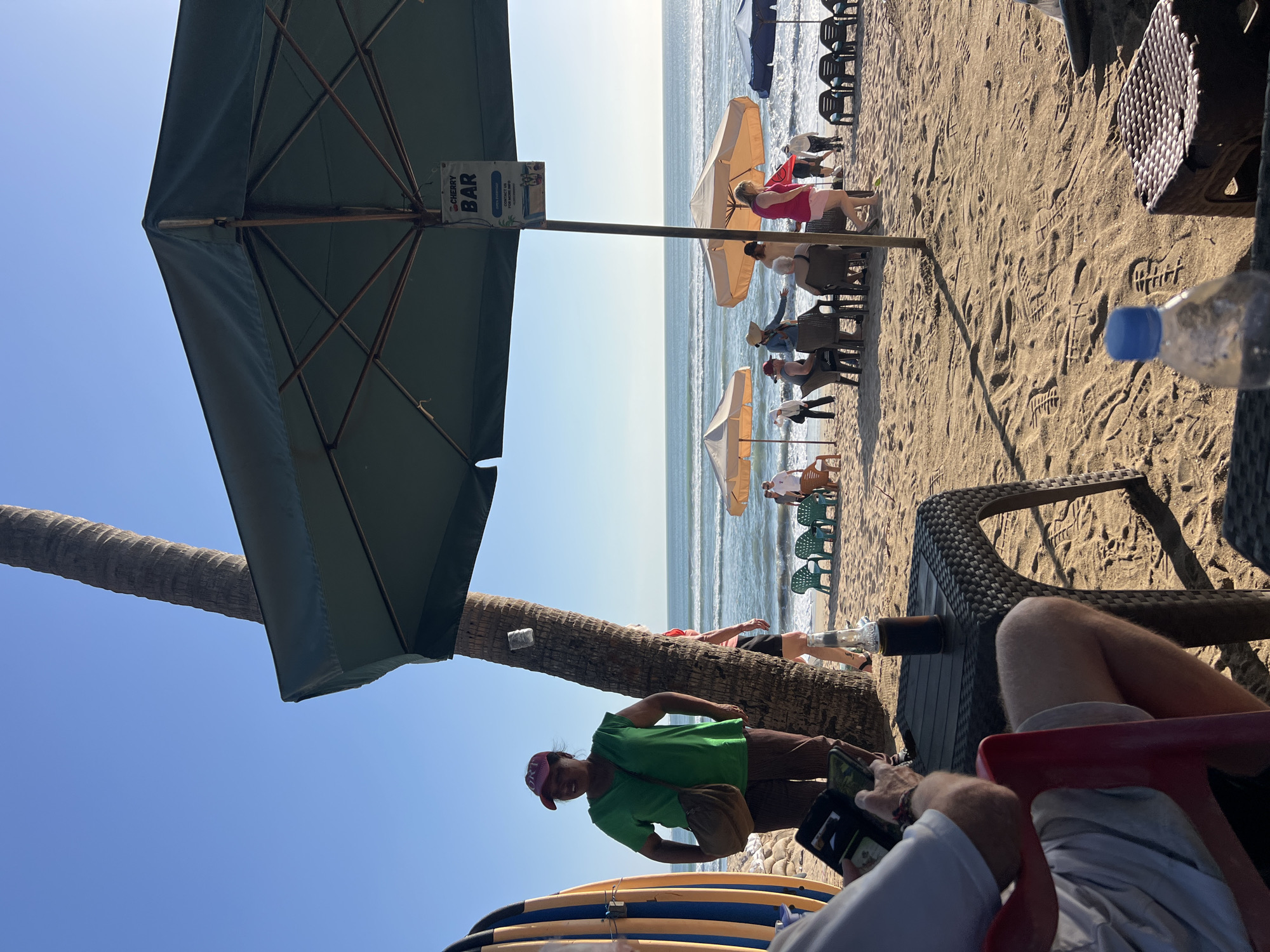 This image captures a sunny day at a beach in Kuta, Indonesia. A large green umbrella, likely for a beach bar, dominates the foreground and casts a shadow on the sand. Beneath the umbrella, a sign reads "CHERRY BAR". Several beach umbrellas and chairs are scattered across the sandy beach, with people relaxing and enjoying the ocean view. The ocean itself is calm with gentle waves. In the left of the image, a person in a green shirt and a red hat stands near a palm tree and surfboards. A table in the foreground holds a bottle and a phone. The scene suggests a relaxed atmosphere at a popular tourist spot.