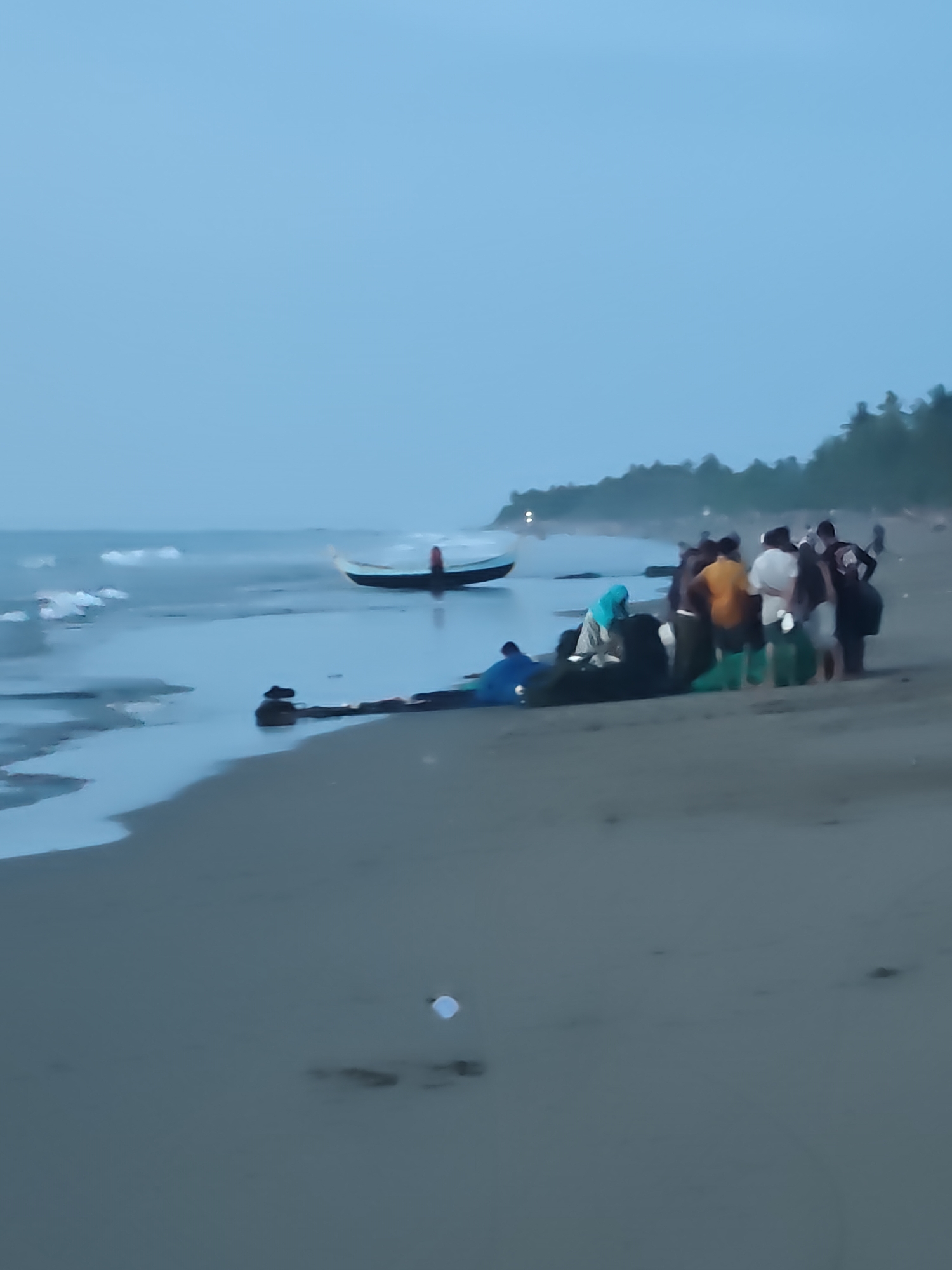 A group of people are gathered on a sandy beach in Curehtunong, Indonesia, near the shoreline. The sky is overcast and the sea is calm with small waves. A traditional fishing boat is beached on the sand, and several individuals are either standing around it or interacting with it. Some of the people are wearing light-colored clothing, while others are in darker attire. The environment suggests a coastal setting with a line of trees visible in the distance. The overall scene appears to be a moment of activity related to the fishing boat on the beach.