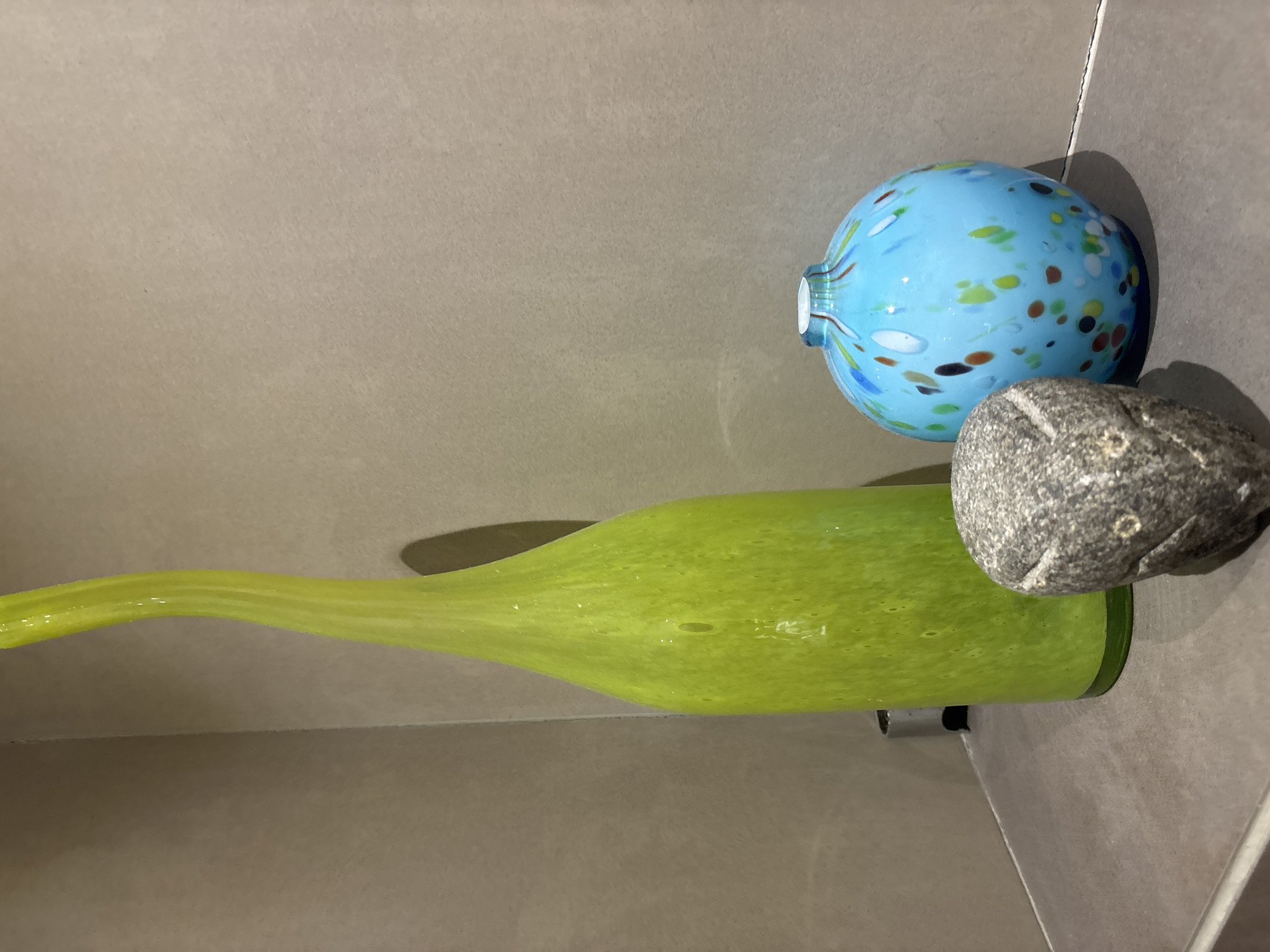 The image shows three decorative objects on a shelf against a tiled wall. On the left is a tall, slender lime green glass vase with a curved neck. To its right is a round, light blue glass vase with multi-colored speckles. In front of the vases is a small, gray, carved stone object that resembles an owl. The objects are placed on a dark gray shelf, and the background consists of light brown textured tiles. There are no people or animals present. The scene is indoors, and based on the modern aesthetic of the objects and tiles, it is plausible this could be in a home, hotel, or shop in Dubai, United Arab Emirates, known for its contemporary design and luxurious interiors.
