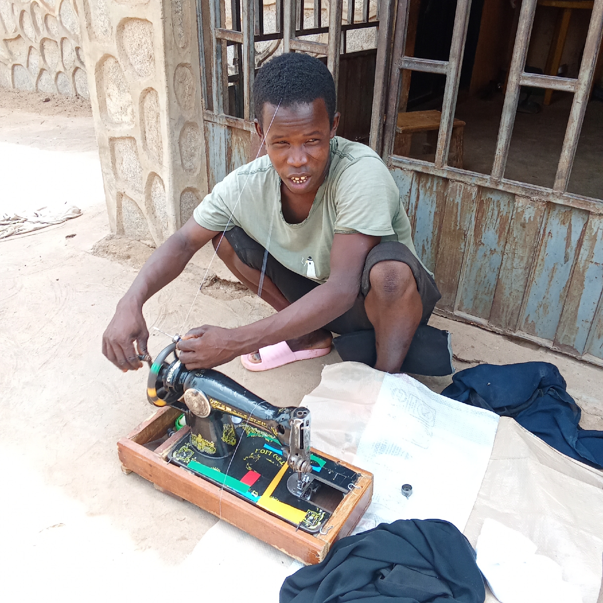 The image depicts a person squatting outdoors in what appears to be a street in Akwanga, Nigeria. The person is seated near a vintage, black sewing machine, which rests on a wooden base. The person is holding a thread and adjusting a part of the sewing machine. The surroundings include stone building structures and a metal gate. Fabric and other sewing-related items are on the ground near the sewing machine. The person is wearing casual clothing, including a t-shirt and shorts. A pink sandal is visible near the person. The ground appears to be a concrete or dirt surface. A chair is inside the building that is behind the gate.