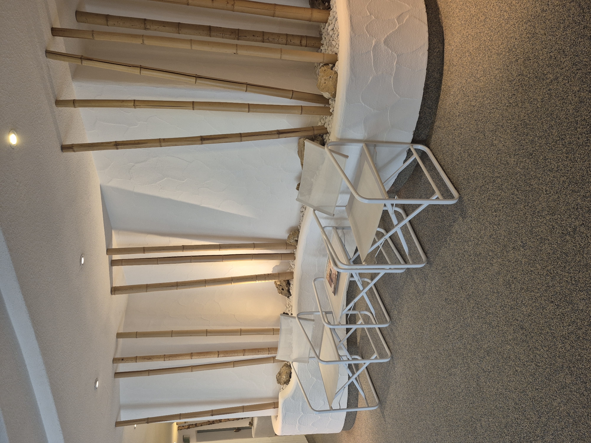 The image shows an indoor seating area with a curved wall decorated with bamboo poles and white, textured plaster. Two white folding chairs with beige canvas seats and backs are positioned in front of the wall, with a small white side table between them. A magazine rests on the table. The floor is covered with speckled grey carpet. The lighting appears to be artificial, with spotlights in the ceiling casting shadows. The scene is devoid of people. The location is identified as the waiting area of the Medical Center St. Josef in Schwäbisch Gmünd, Germany.