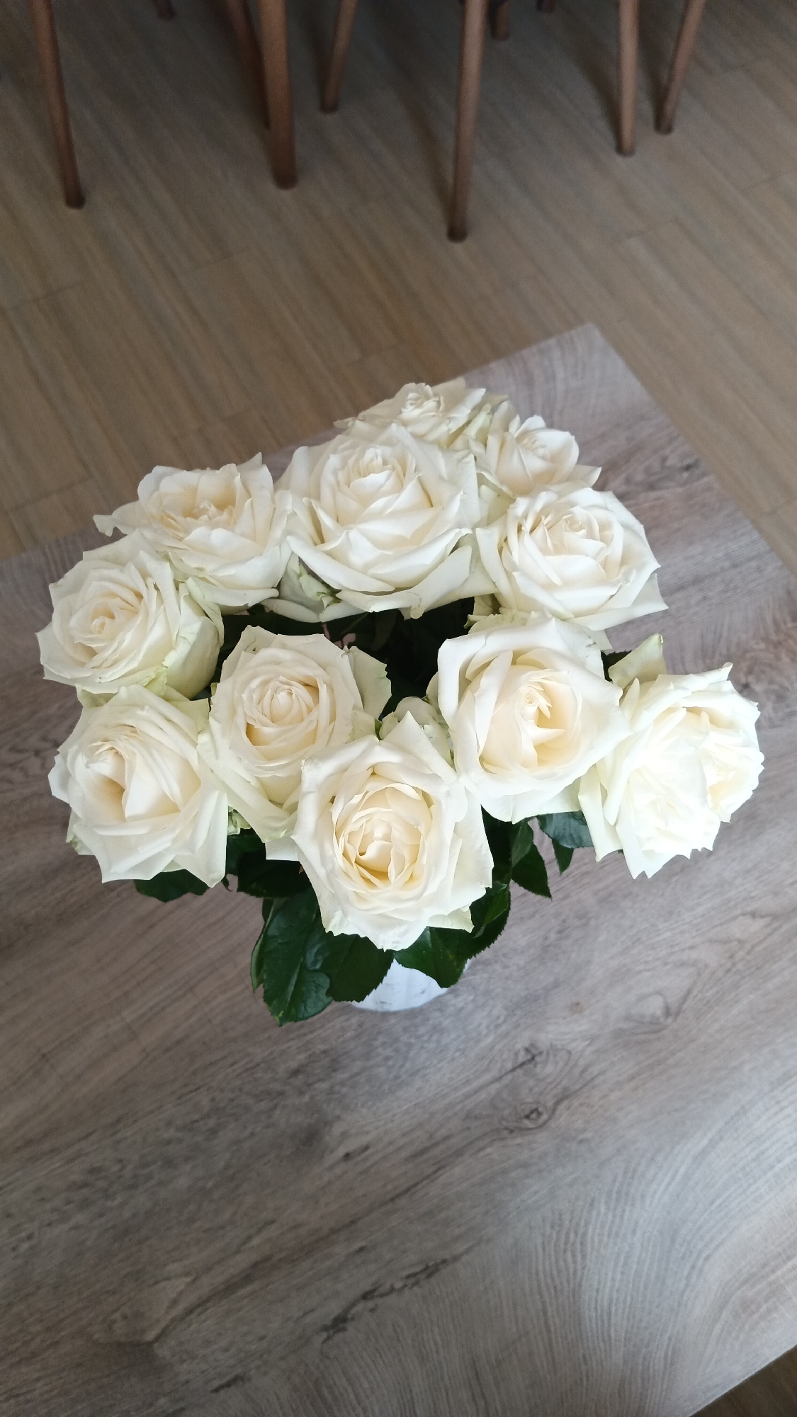 A bouquet of white roses is displayed in a white vase on a wooden table. The table has a light brown wood grain pattern. In the background, the legs of wooden chairs are visible, suggesting the scene is indoors, possibly in a home or a cafe. The lighting is natural, casting soft shadows. The image does not contain any information about the city of Ashtarak, Armenia.