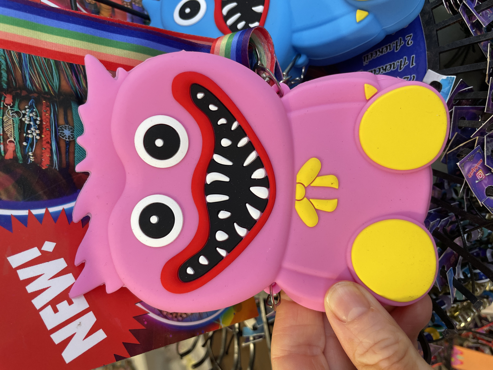The image shows a pink, fuzzy character toy with big white eyes, red lips, and sharp, black and white teeth. It has a yellow bow on its chest and yellow circular feet. The toy is held by a person's hand, which is visible on the left side of the frame. Behind the toy, there are other toys and a display rack. The background includes a "NEW!" sign in red and a colorful rainbow-striped ribbon. The context of Sochi, Russia, is not evident from the visual information provided in the image.