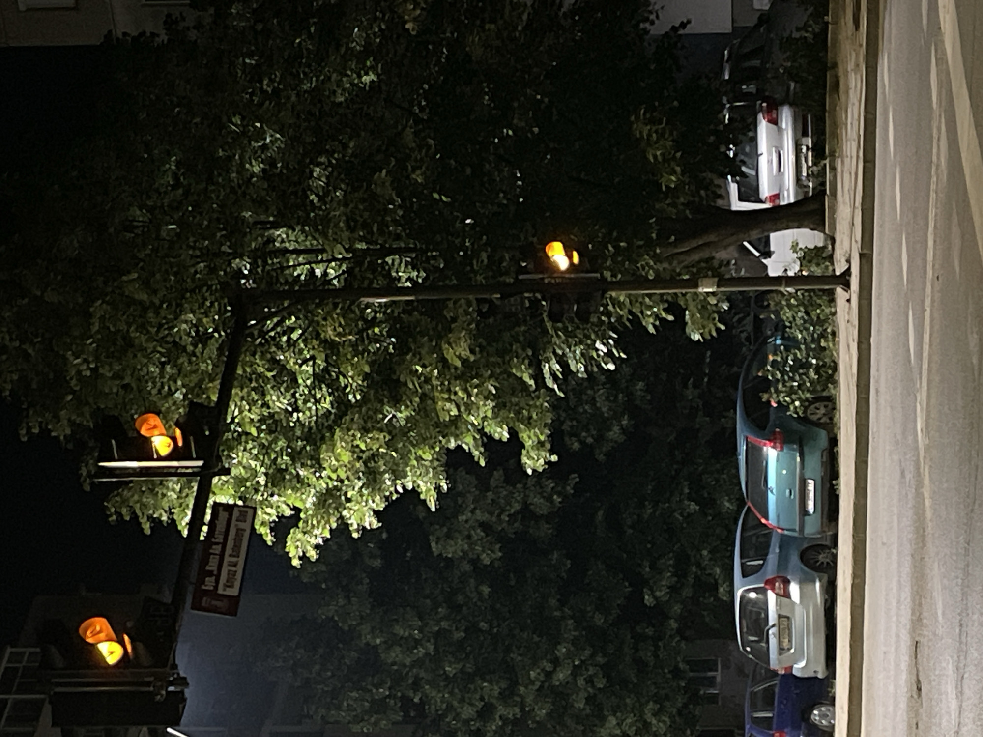 The image displays a nighttime street scene, rotated 90 degrees clockwise, predominantly featuring a large, dark-leafed tree that occupies much of the frame. The tree's foliage is partially illuminated by artificial light.
Multiple traffic light heads are visible, all illuminated with an amber signal, suggesting a yellow phase or a flashing cautionary state. One set of traffic lights is mounted on a pole that also holds a street sign. The sign bears text in what appears to be Greek; the upper line is partially decipherable as "Evg. Kira An. Stefa...", and the lower line reads "Kyra An. Rotem...". These names suggest the location is in a Greek-speaking country.
Along the right side of the street (after mental rotation to correct orientation), several cars are parked parallel to a curb. A silver hatchback is prominently visible, with a light blue hatchback parked directly behind it, and a dark blue car partially seen further back. Another silver car is visible parked on the right, partially obscured by the tree. A building is faintly discernible in the background behind the tree.
The overall setting is an urban or suburban street at night, with no human activity or specific events taking place. The time of day is clearly night, evidenced by the darkness and the reliance on artificial light sources. No specific weather conditions are indicated, and no emotions are conveyed by the static scene.